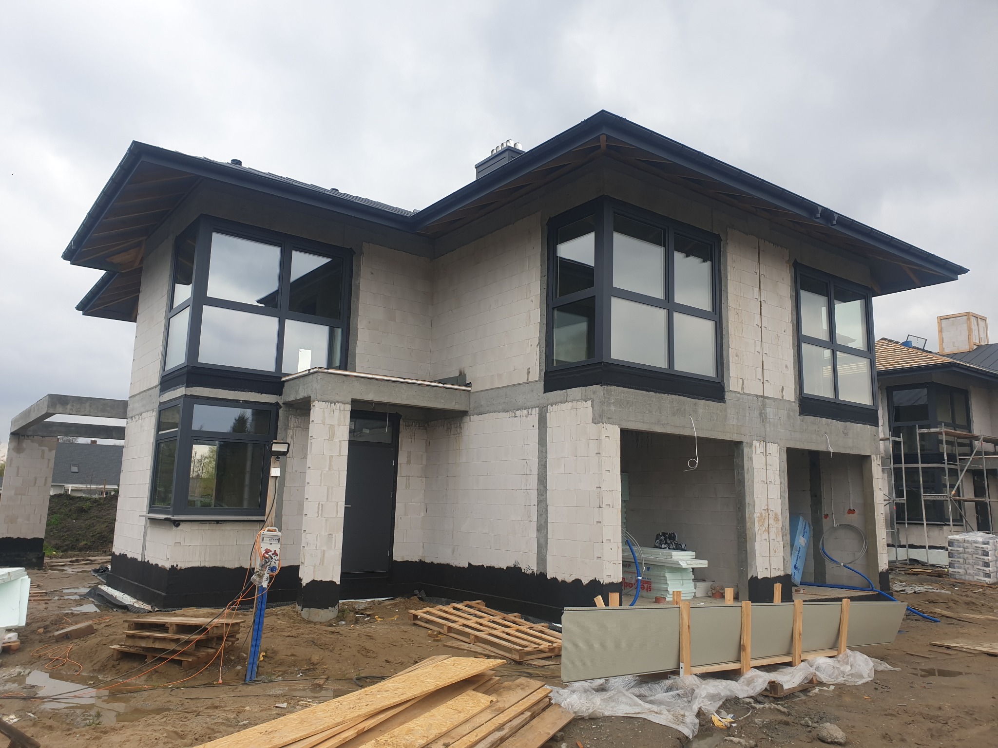 Surowy stan dwupiętrowego domu z białego betonu komórkowego z zamontowanymi ciemnymi oknami i czarnym dachem, widoczne palety, deski i materiały budowlane na zewnątrz.