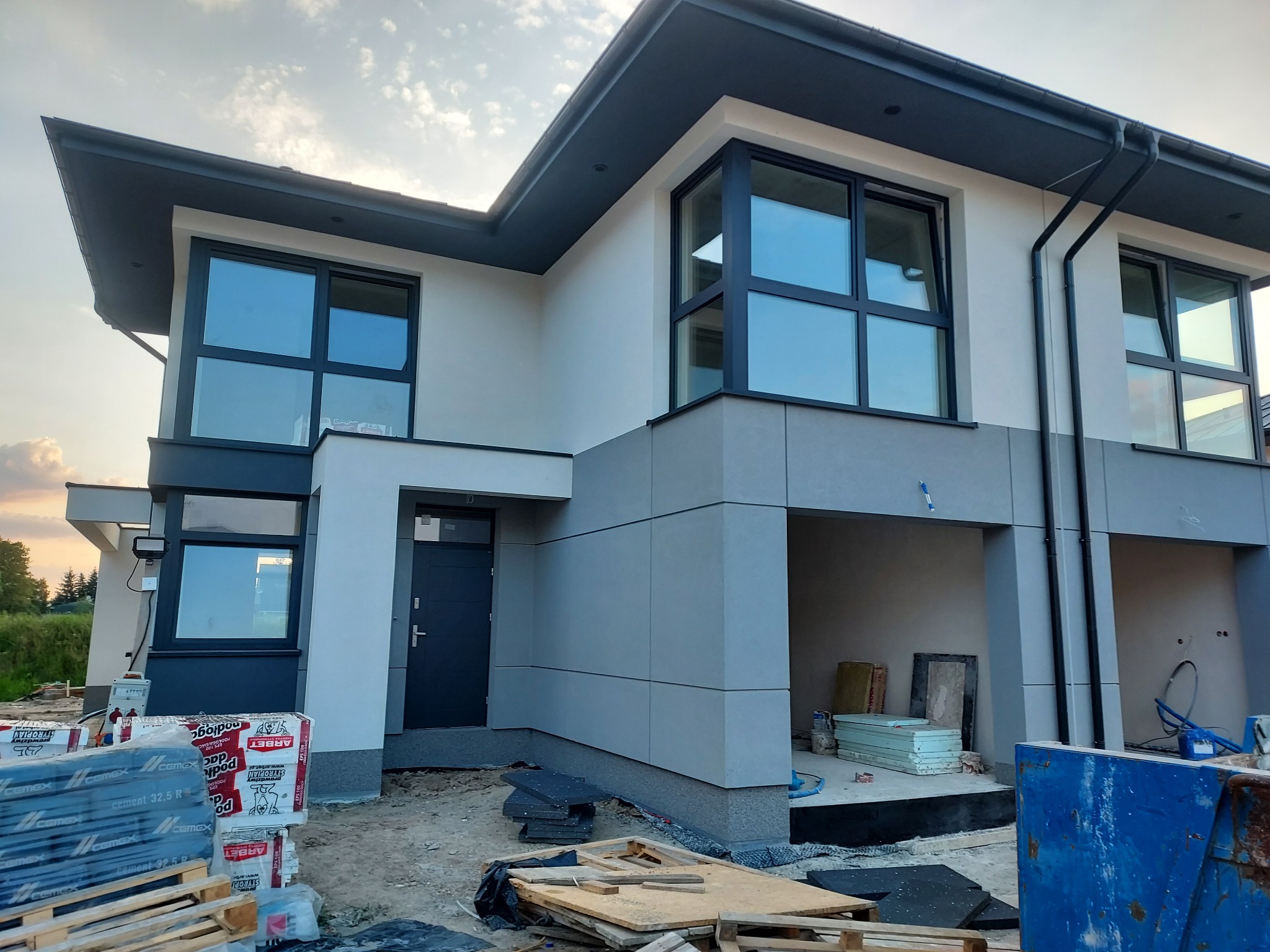 Nowoczesny dom w trakcie budowy, z widoczną elewacją w odcieniach szarości i bieli, duże okna z czarnymi ramami, materiały budowlane na paletach przed budynkiem, niebieski kontener. Widoczne...