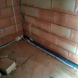 ELTIM - Usługi Elektryczne Wacław Dawid - Wnętrze budynku w trakcie prac instalacyjnych: kable w peszlach ułożone wzdłuż ściany z czerwonej cegły dziurawki, zabezpieczone pianką montażową.