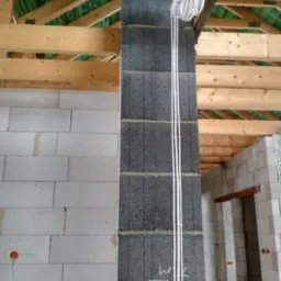 Wewnętrzna instalacja elektryczna w surowym budynku z szarych bloczków, kable przymocowane do ściany, widoczne drewniane elementy konstrukcji dachu.