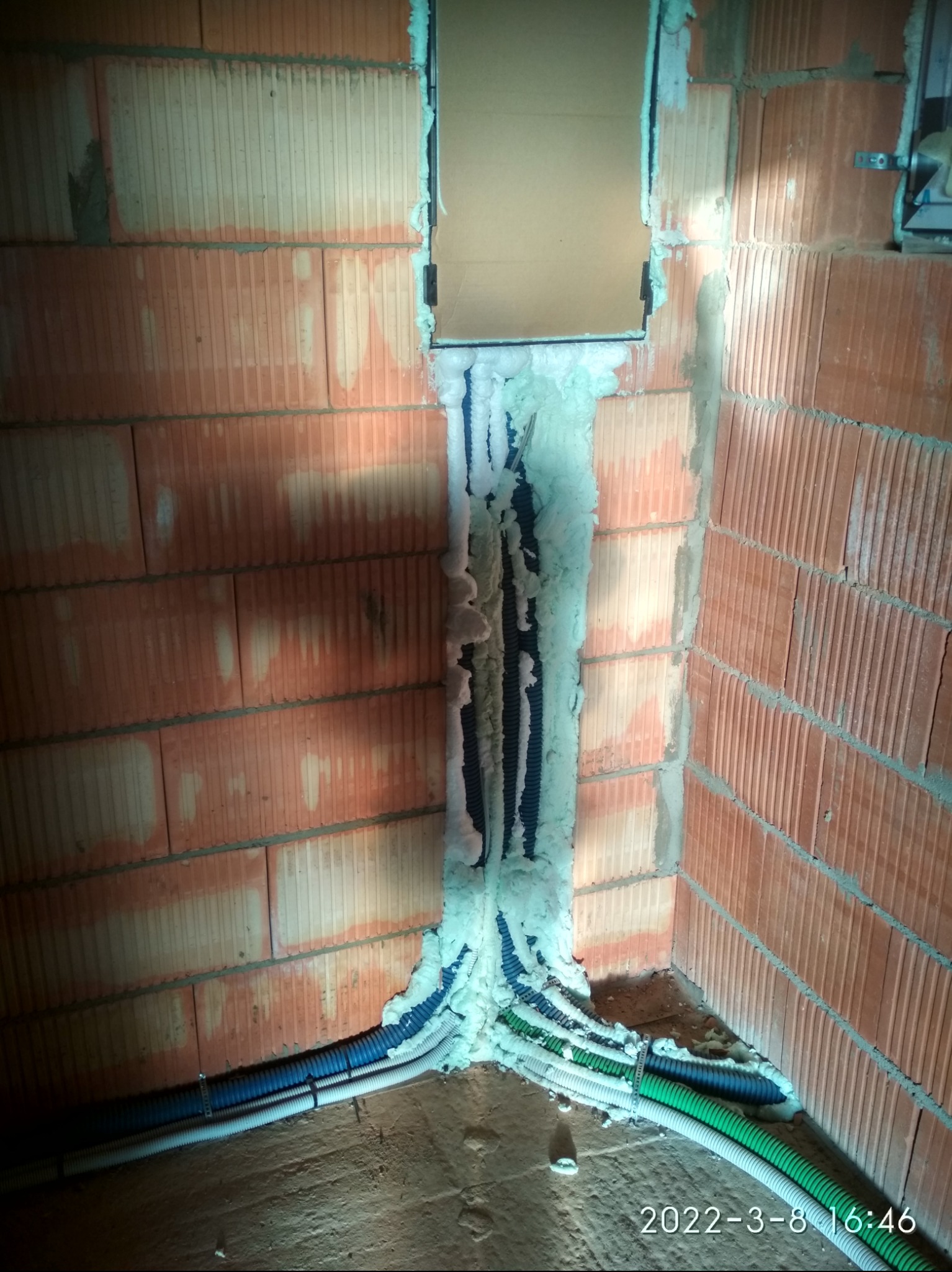 Wewnętrzny róg z cegły z pionowym kanałem wypełnionym pianką montażową, przez który przechodzą rury instalacyjne w kolorach: niebieskim, zielonym i białym.