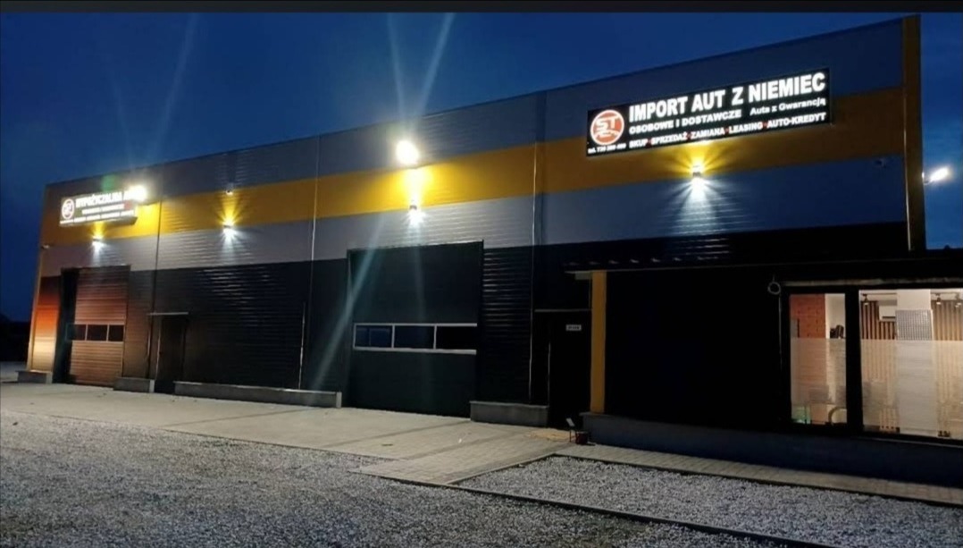 Fasada budynku firmy 'Import Aut z Niemiec' oświetlona lampami zewnętrznymi, z widocznymi bramami garażowymi i oknami, sfotografowana nocą.