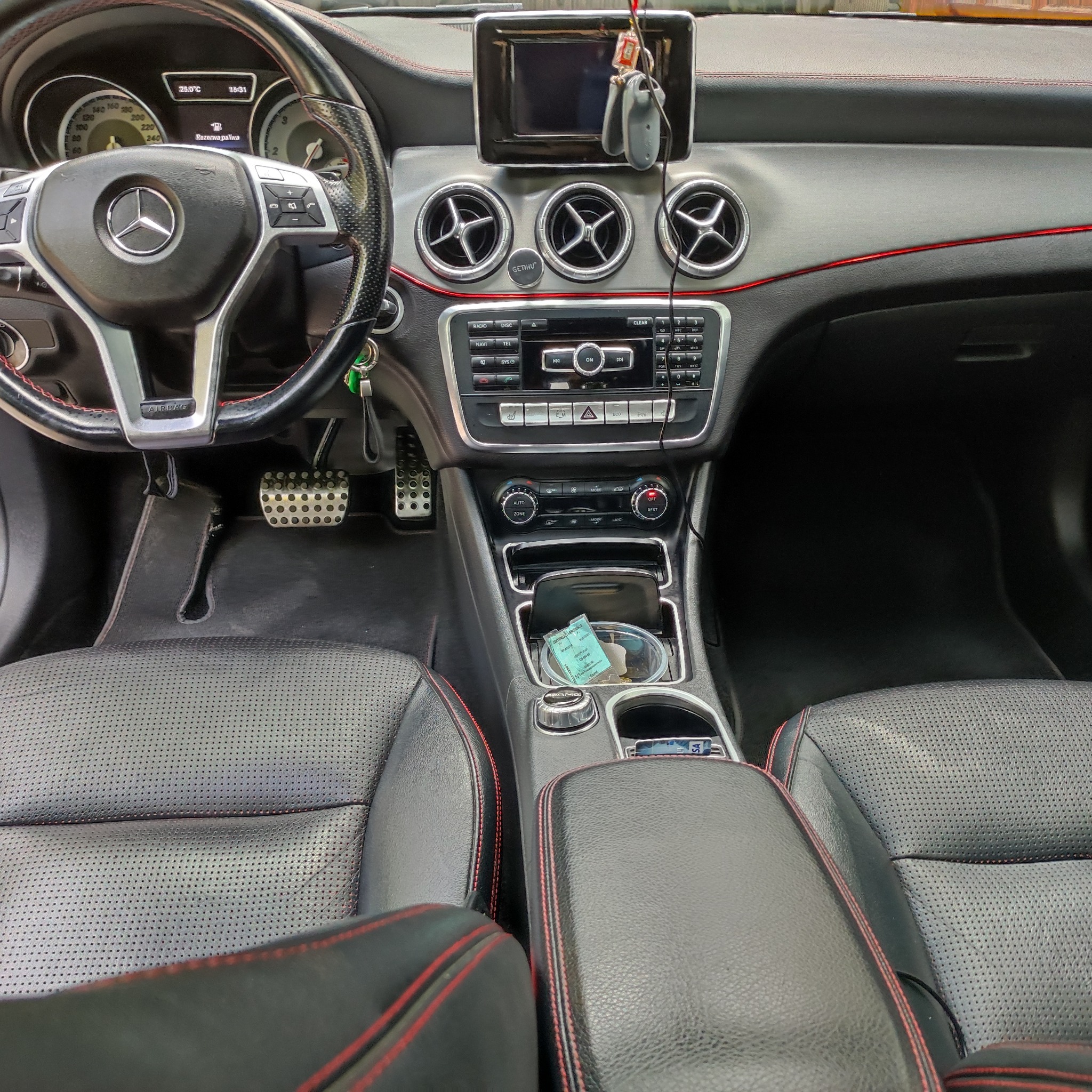 Wnętrze samochodu marki Mercedes, widok na deskę rozdzielczą, kierownicę z logo, skórzane fotele z czerwonymi przeszyciami oraz panel sterowania klimatyzacją i radiem.