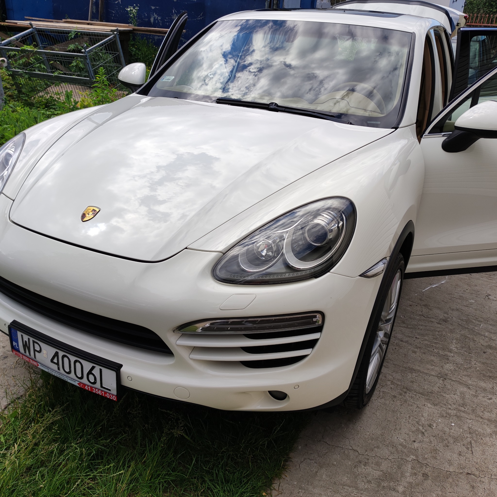 Biały samochód marki Porsche z otwartymi drzwiami, zaparkowany na betonowej nawierzchni obok trawy.