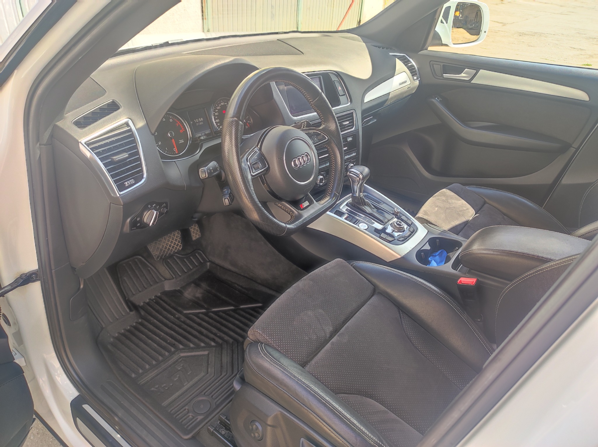 Wnętrze białego Audi Q5 z czarną skórzaną tapicerką, widoczne gumowe dywaniki i deska rozdzielcza z logo Audi na kierownicy.