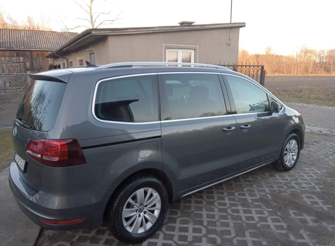 Szary minivan Volkswagen Sharan zaparkowany na betonowej kostce brukowej, z widocznym budynkiem w tle i drzewami bez liści.