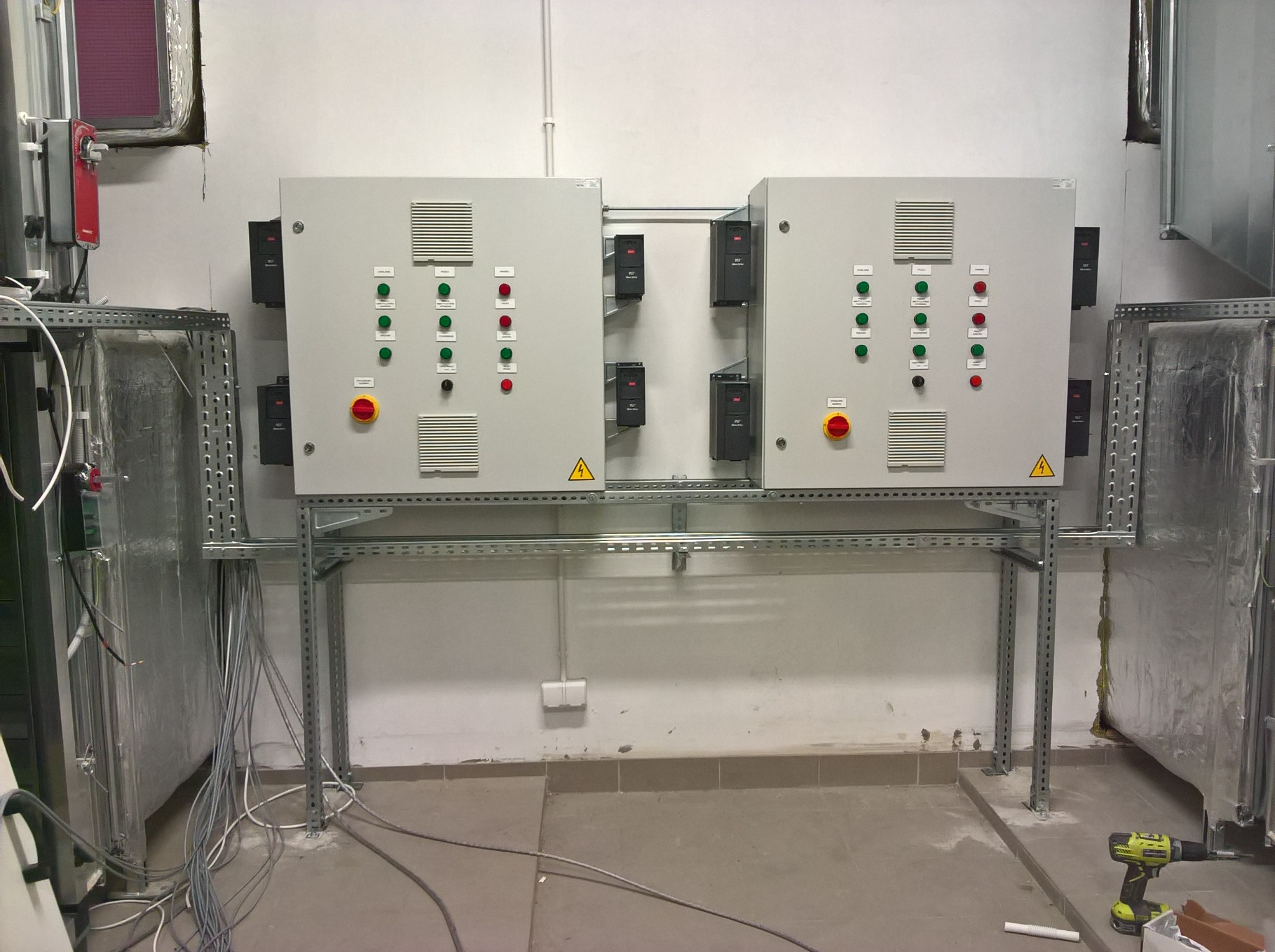 Dwa szare panele sterowania elektrycznego zamontowane na metalowej konstrukcji w pomieszczeniu technicznym. Widoczne kable, wiertarka akumulatorowa i elementy wentylacji.