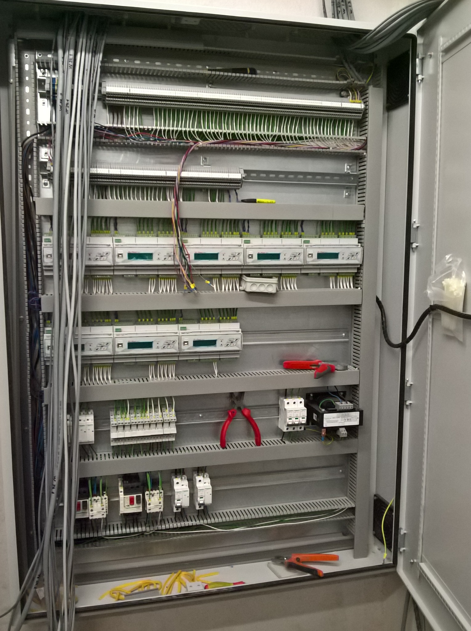 Otwarta szafa sterownicza z licznymi przewodami, bezpiecznikami i urządzeniami pomiarowymi. Widoczne narzędzia: kombinerki z czerwonymi i pomarańczowymi uchwytami.