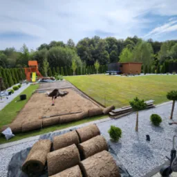 Rozwinięte rolki trawy na przygotowanym podłożu, widoczne narzędzia i fragment trawnika, w tle drzewa i plac zabaw.