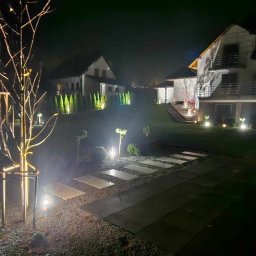 Firma Usługowa 2P inż. Paweł Puto - Oświetlony ogród nocą z podświetlonymi drzewami, ścieżką z płyt i budynkiem mieszkalnym w tle.