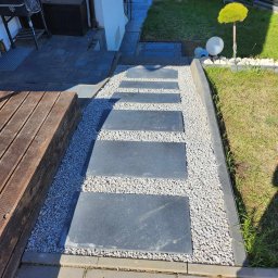 Firma Usługowa 2P inż. Paweł Puto - Ścieżka z ciemnych płyt kamiennych, otoczona białym żwirem, prowadząca przez trawnik w ogrodzie, obok drewnianego tarasu i domu.