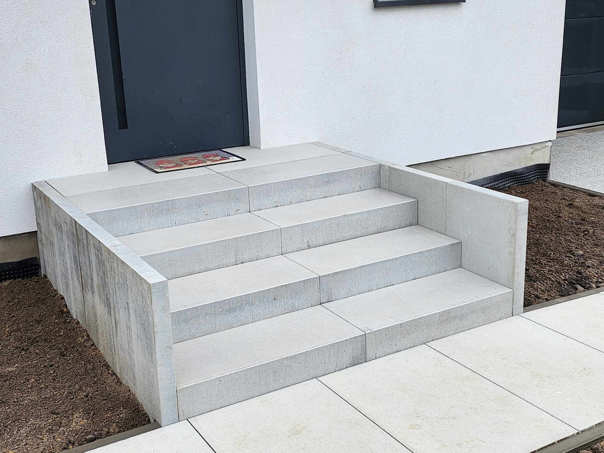 Nowoczesne, jasnoszare schody wejściowe z kostki betonowej prowadzące do ciemnych drzwi, z wycieraczką z motywem krasnali, obok świeżo obsypana ziemia.