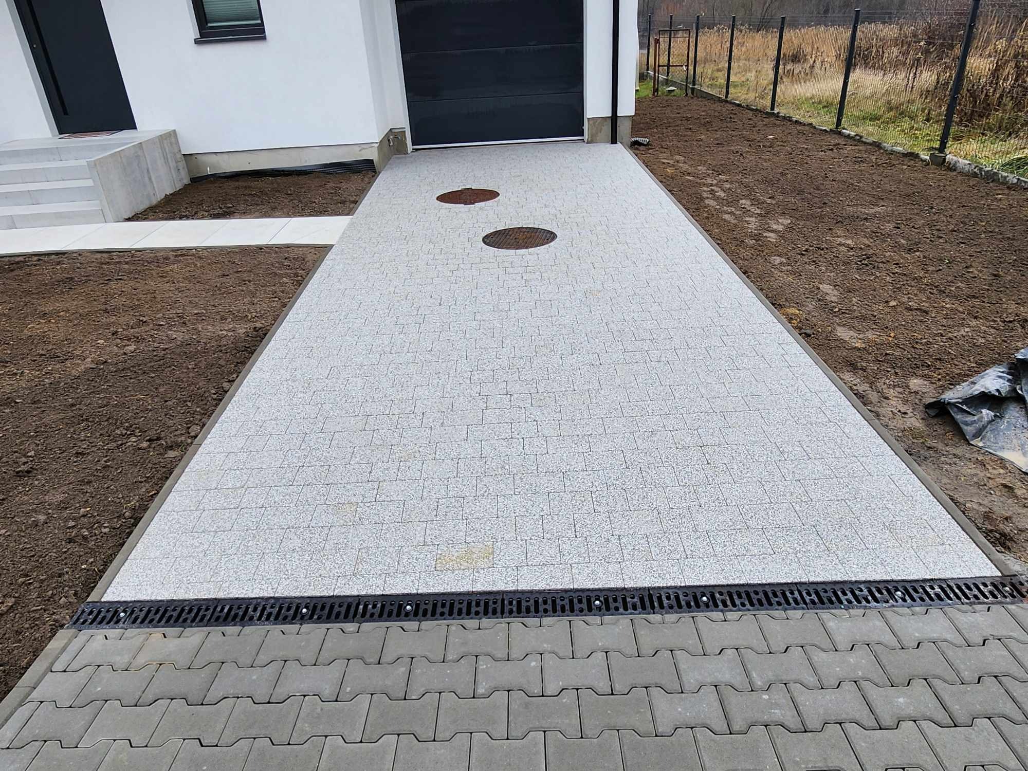 Nowa, jasnoszara nawierzchnia z kostki przed garażem i domem, z dwoma okrągłymi włazami i czarnym odpływem liniowym przy krawędzi, obok świeżo przygotowany teren pod ogród.