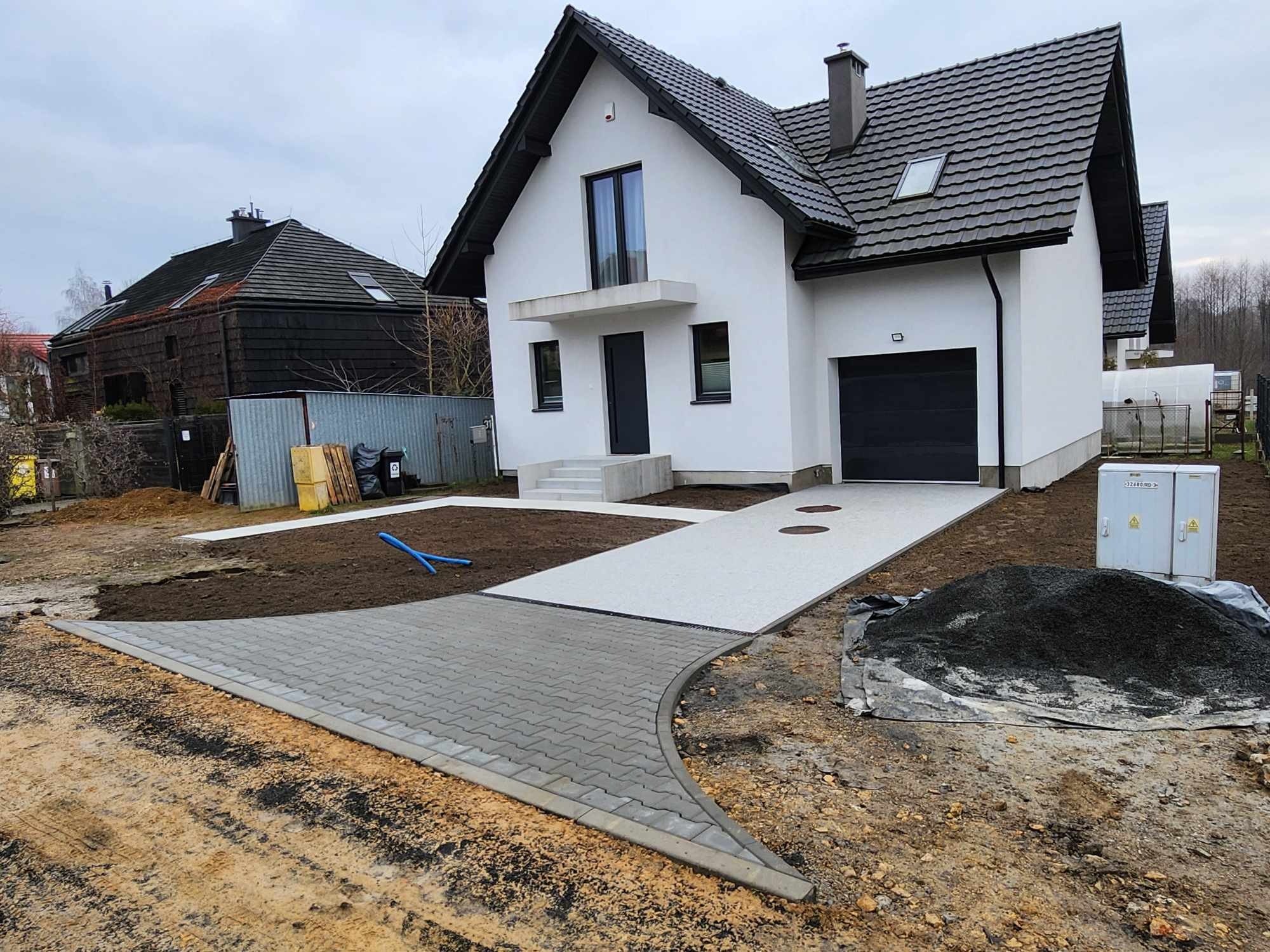 Nowoczesny dom jednorodzinny z szarą kostką brukową na podjeździe, świeżo ułożoną na łuku, z widocznym przygotowanym terenem pod trawnik i stertą ziemi obok.