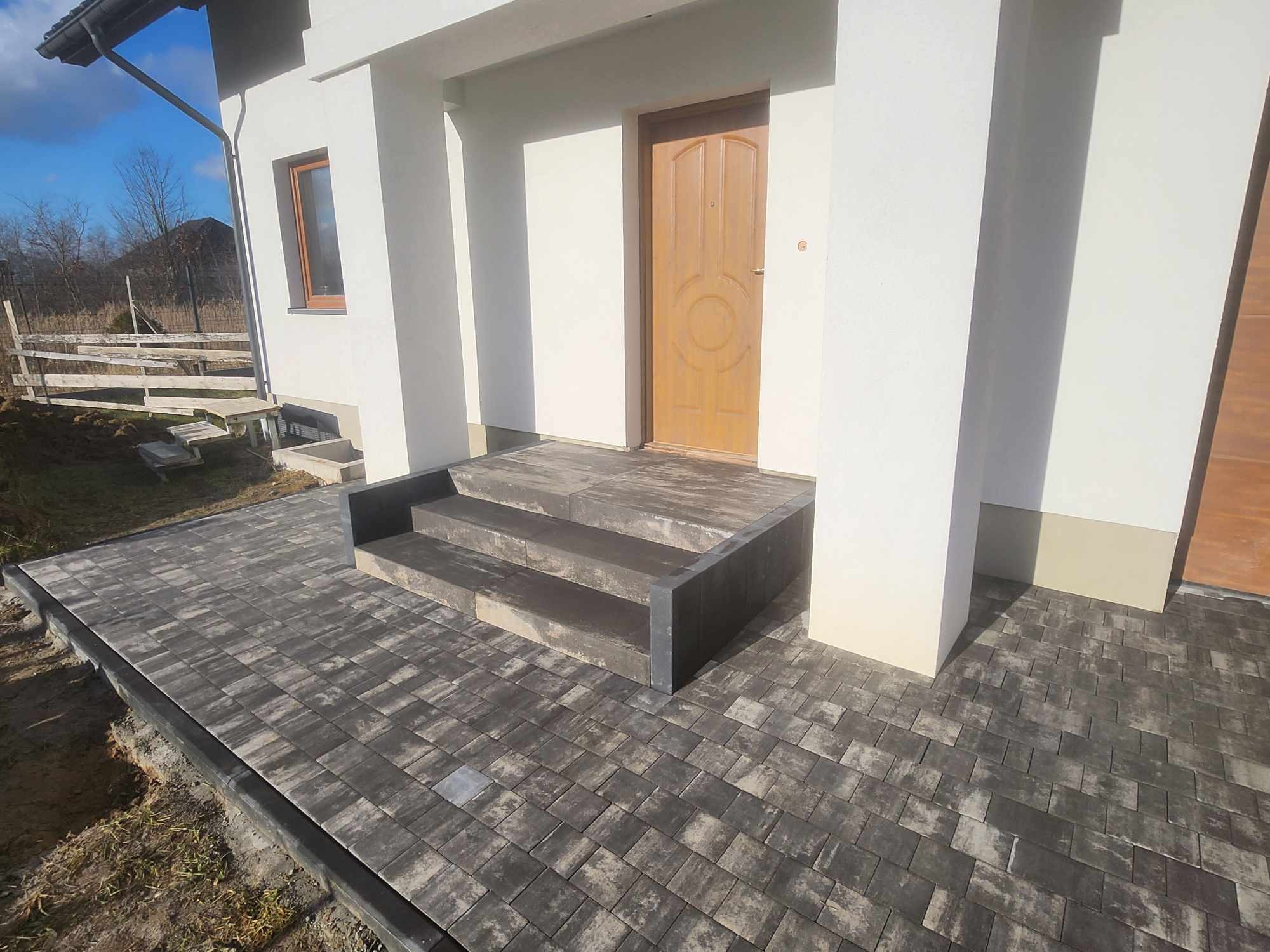 Nowo ułożona, szara kostka brukowa prowadząca do wejścia domu z trzema schodami, widoczne obrzeża i surowy teren po bokach.