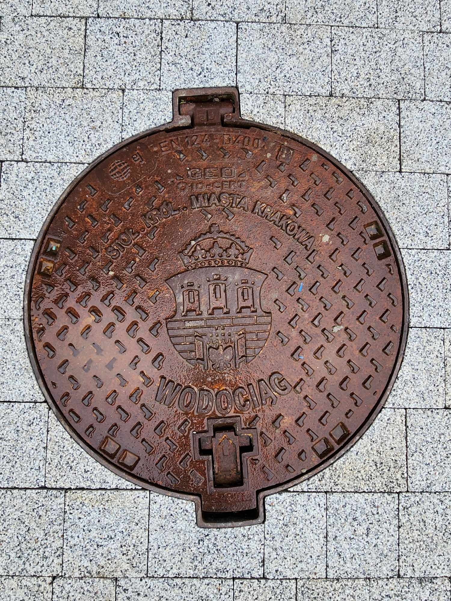 Okrągła, żeliwna studzienka kanalizacyjna z herbem Krakowa, osadzona w szarej kostce brukowej. Widoczne napisy 'WODOCIĄGI' i 'STOŁ. KRÓL. MIASTA KRAKOWA'.