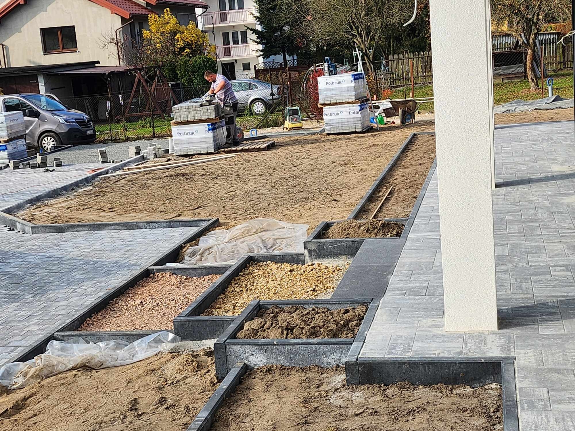 Układanie kostki brukowej: widok na przygotowany teren z wydzielonymi kwaterami wypełnionymi różnym materiałem (piasek, żwir, ziemia) oraz pracownika układającego kostkę brukową Polbruk z palet w tle.