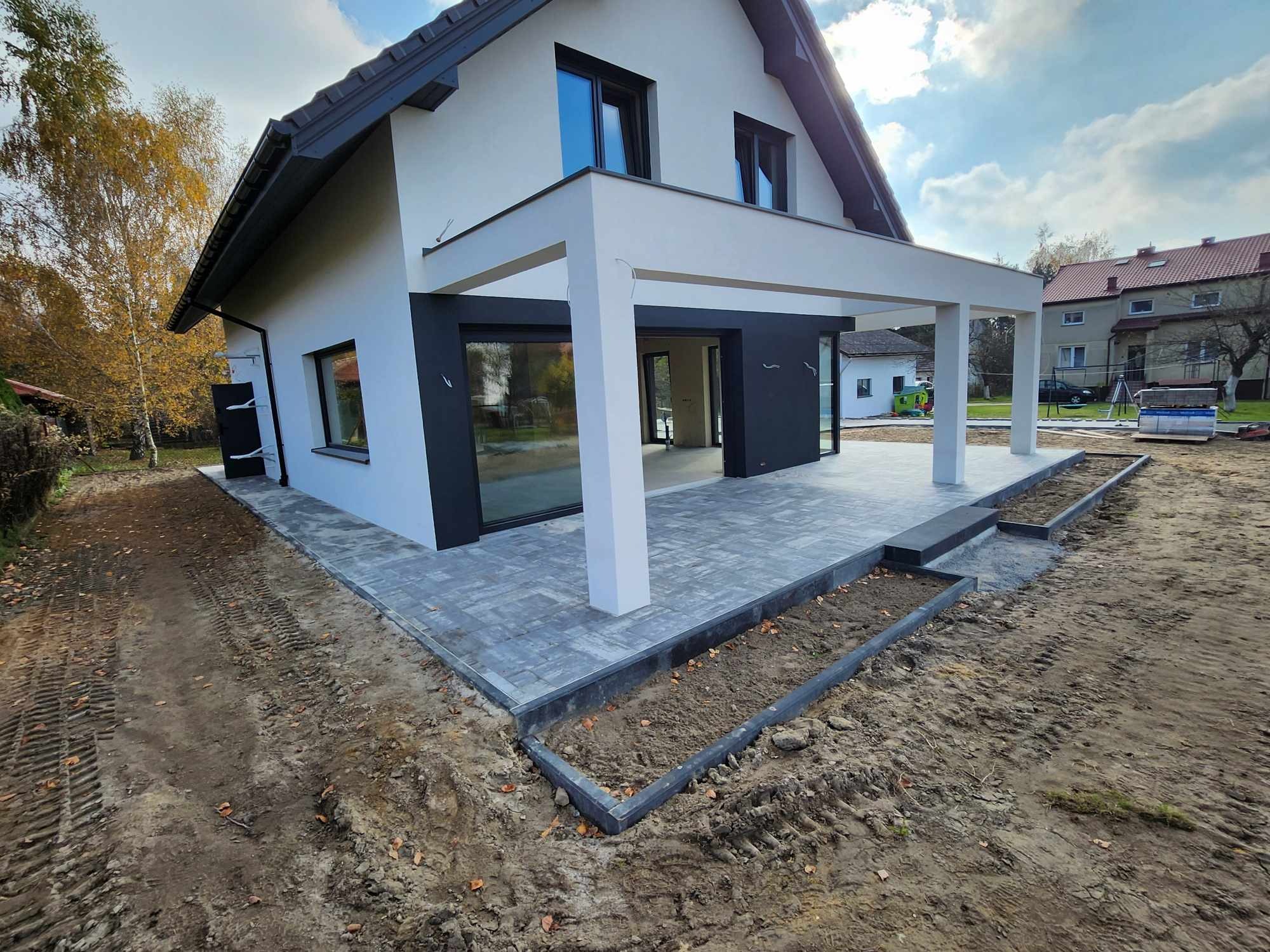 Nowoczesny taras z szarej kostki, obrzeża z ciemnego betonu, biały dom z dużymi oknami, teren wokół w trakcie zagospodarowywania, widoczne ślady opon.