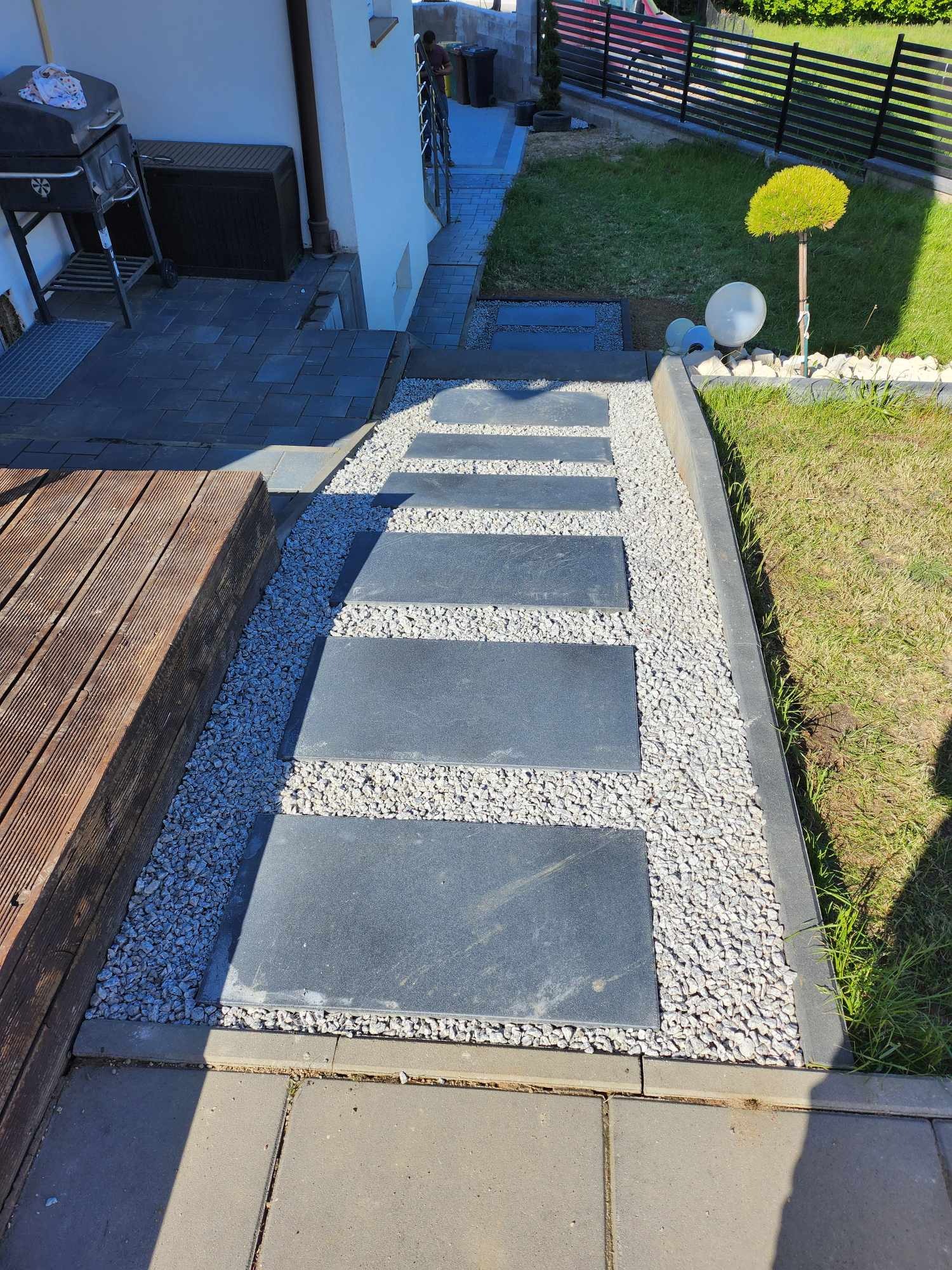 Ścieżka z ciemnych płyt kamiennych, otoczona białym żwirem, prowadząca przez trawnik w ogrodzie, obok drewnianego tarasu i domu.