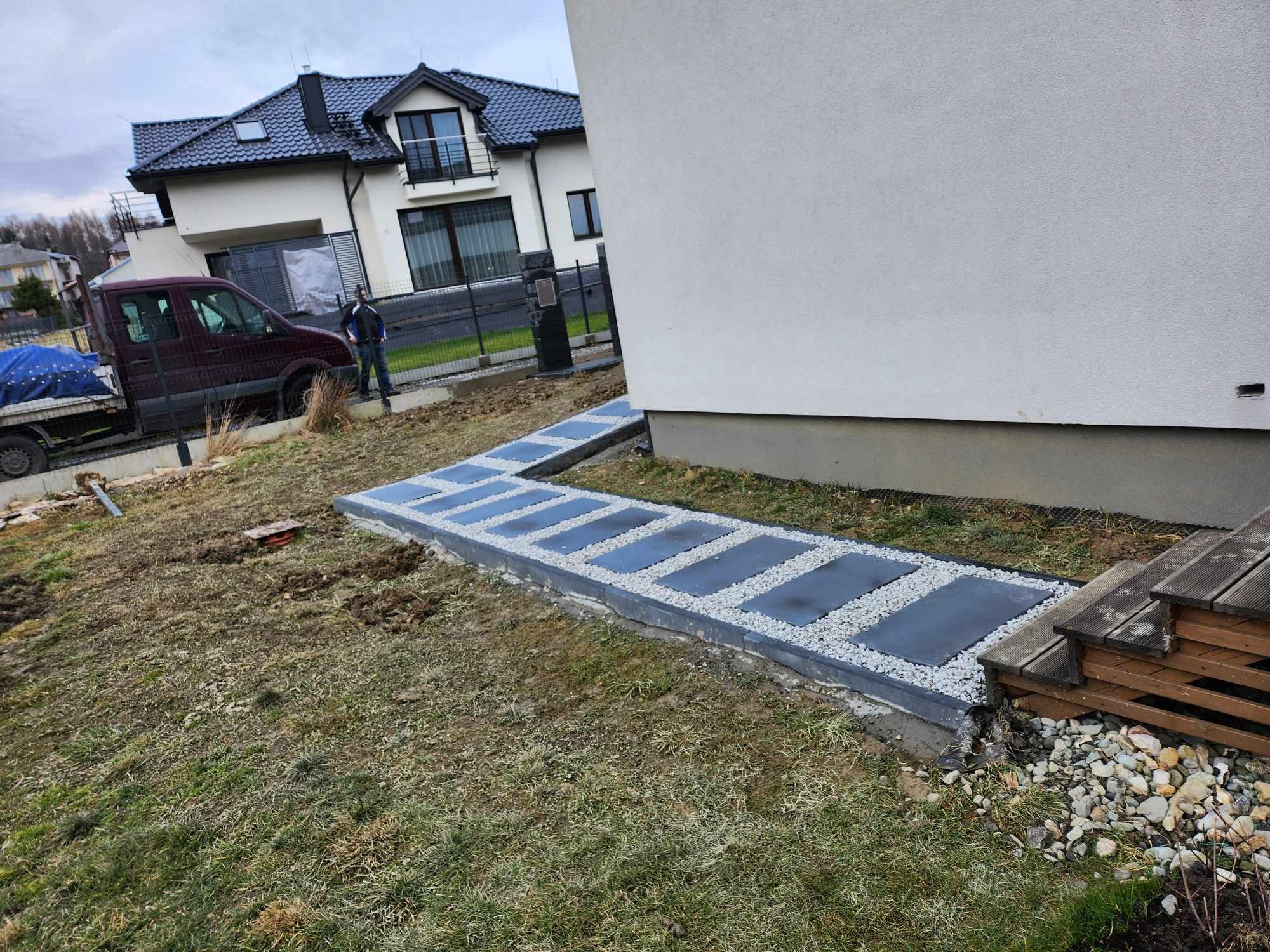 Ścieżka z prostokątnych płyt ułożona w trawie, oddzielona od gruntu drobnym żwirem, prowadząca wzdłuż ściany domu, z widocznym fragmentem drewnianego podestu i samochodem dostawczym w tle.