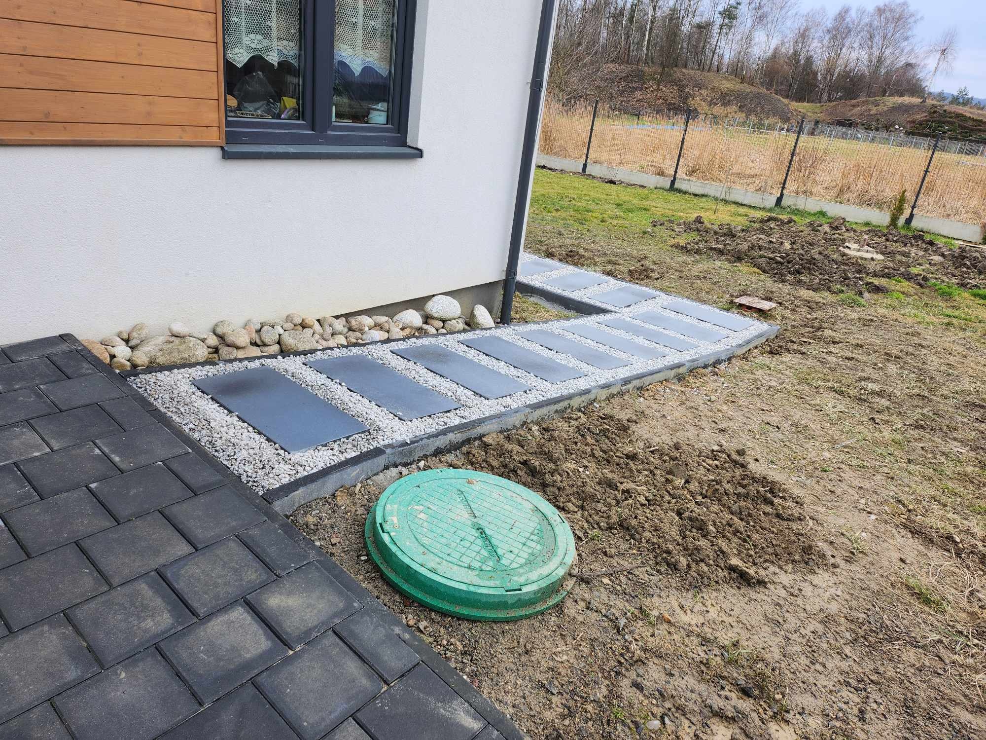 Ścieżka z płyt betonowych oddzielonych drobnym żwirem, prowadząca wzdłuż ściany domu, obok studzienki kanalizacyjnej i kostki brukowej.