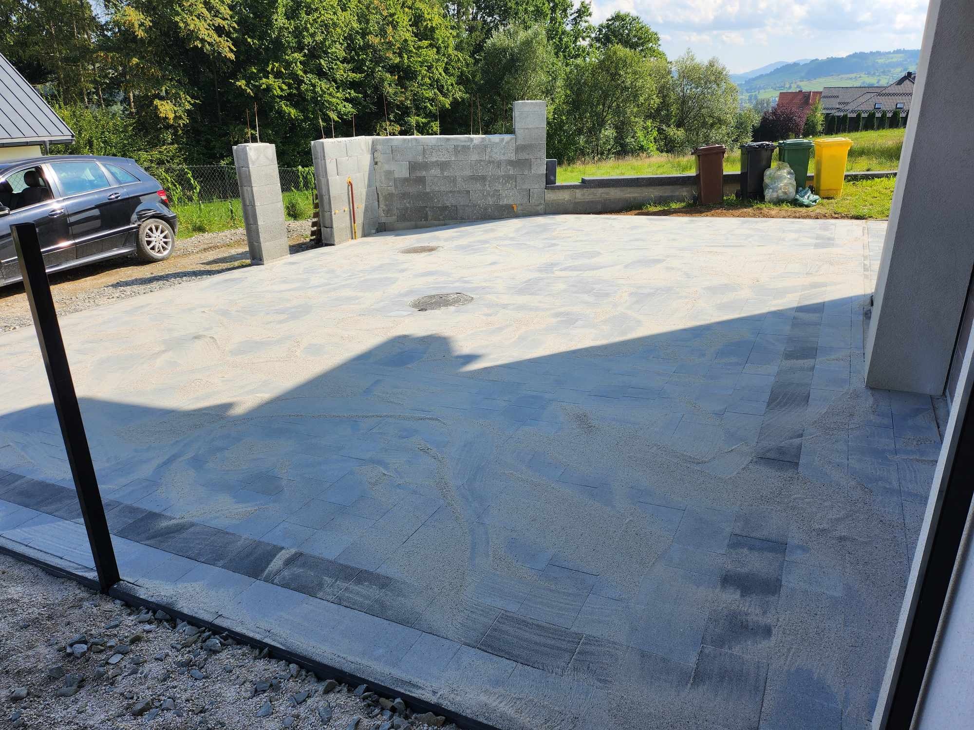 Nowa, szara nawierzchnia z kostki, częściowo zasypana piaskiem, otoczona elementami betonowymi i widokiem na zielony krajobraz z drzewami i wzgórzami w tle.