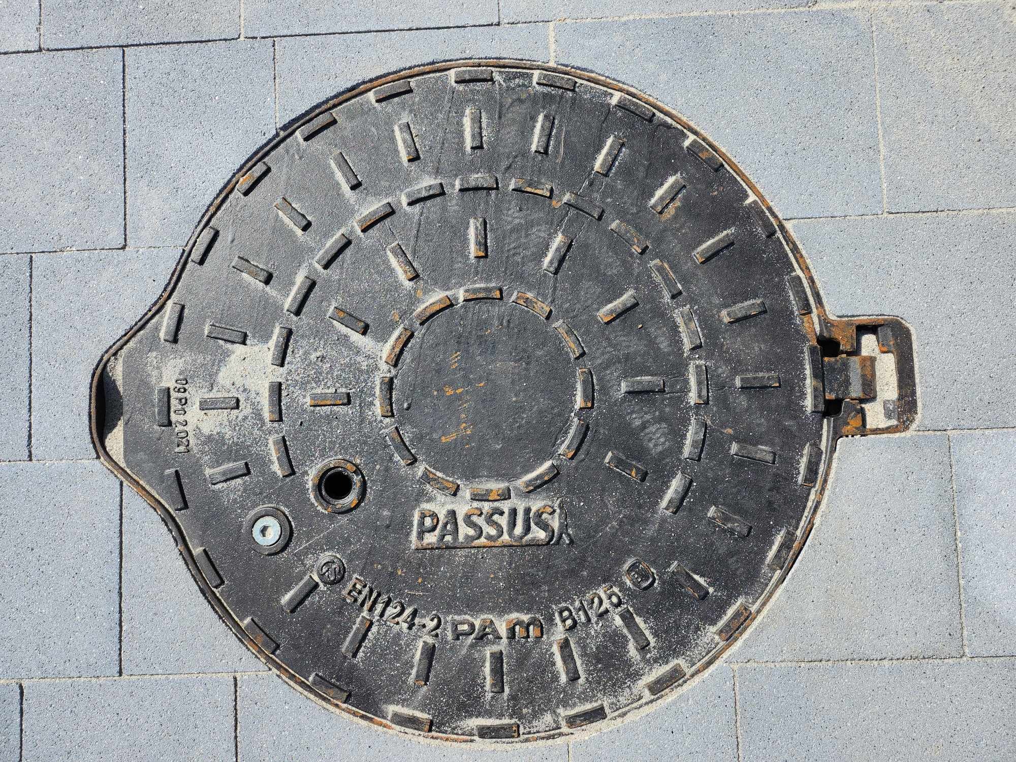 Żeliwna pokrywa włazu kanalizacyjnego firmy PASSUS wkomponowana w szarą kostkę brukową, widoczne oznaczenia normy EN124-2 PAM B125 oraz ślady piasku na powierzchni.