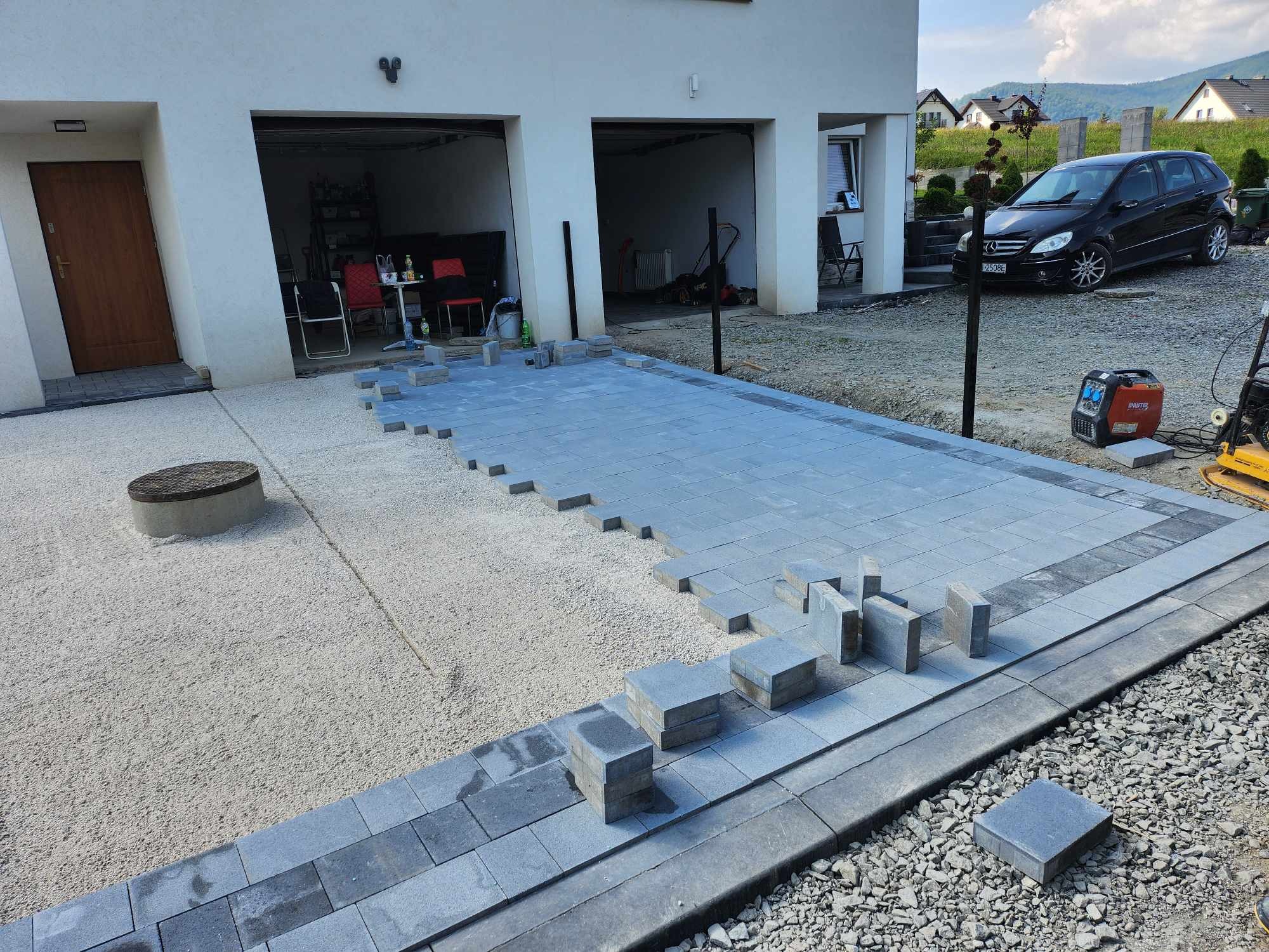 Częściowo ułożona, szara nawierzchnia z kostki przed domem z dwoma garażami, widoczne narzędzia i samochód w tle, w trakcie prac.