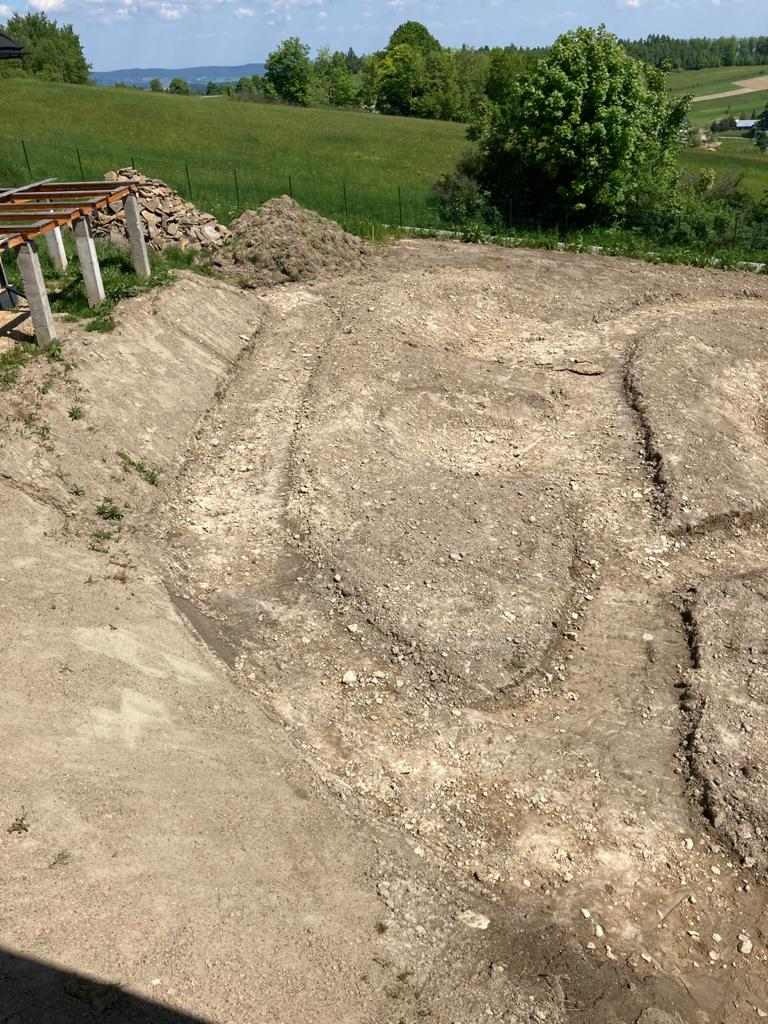 Przygotowany teren pod przyszły ogród z widocznymi śladami po wykopach, stertą ziemi i elementami konstrukcji w tle, na tle zieleni wzgórz.