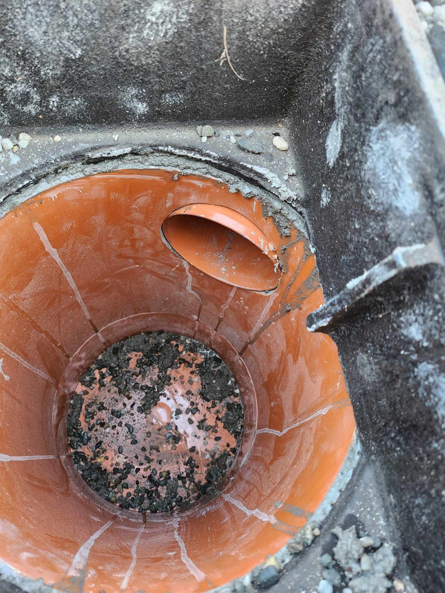 Pomarańczowa studzienka kanalizacyjna z doprowadzoną rurą i wypełniona żwirem w betonowej obudowie.