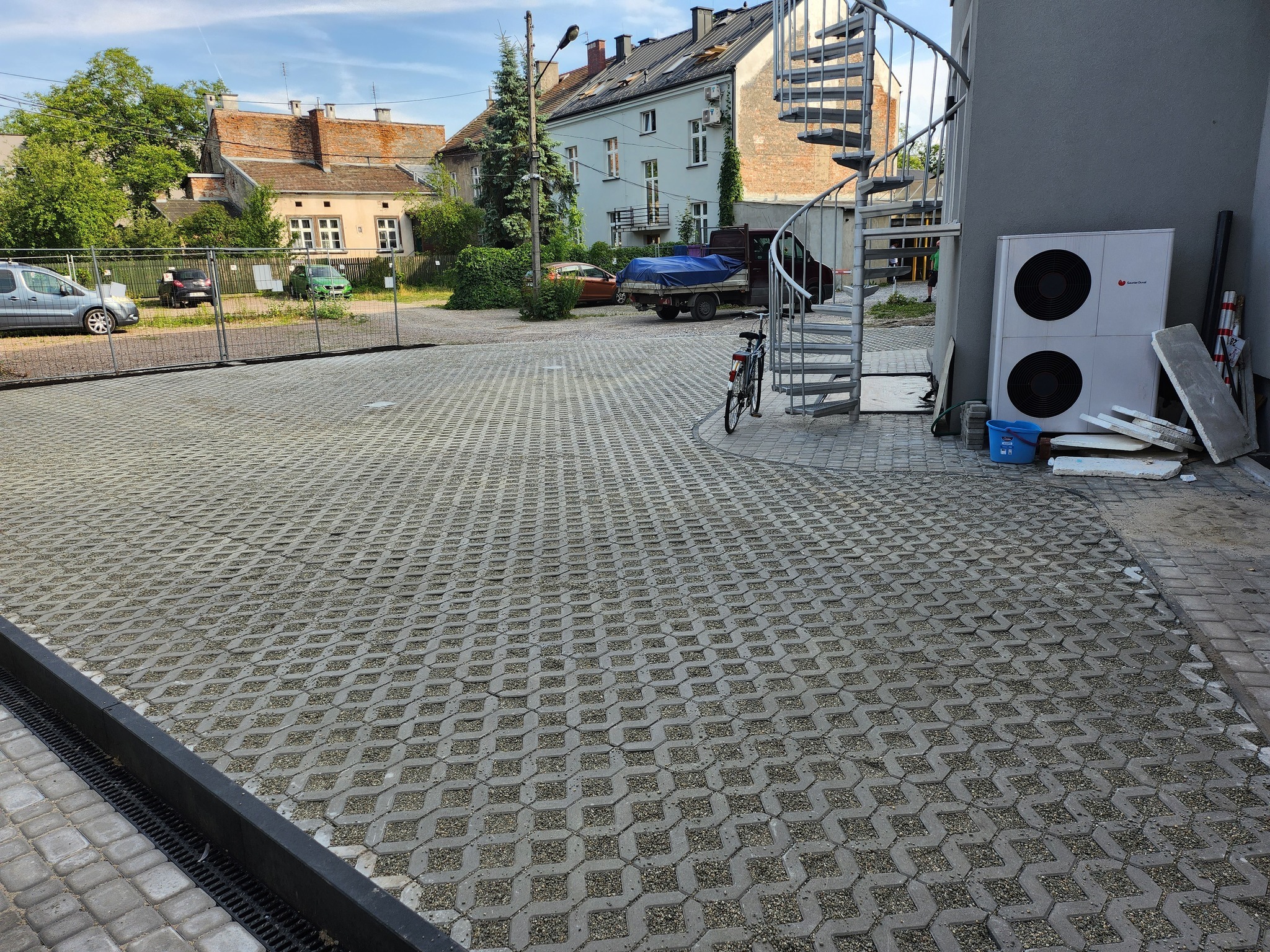 Szeroki widok na parking wyłożony ażurową kostką betonową, z widocznymi elementami otoczenia: budynki mieszkalne, drzewa, rower oparty o spiralne schody zewnętrzne, oraz klimatyzator przy ścianie...