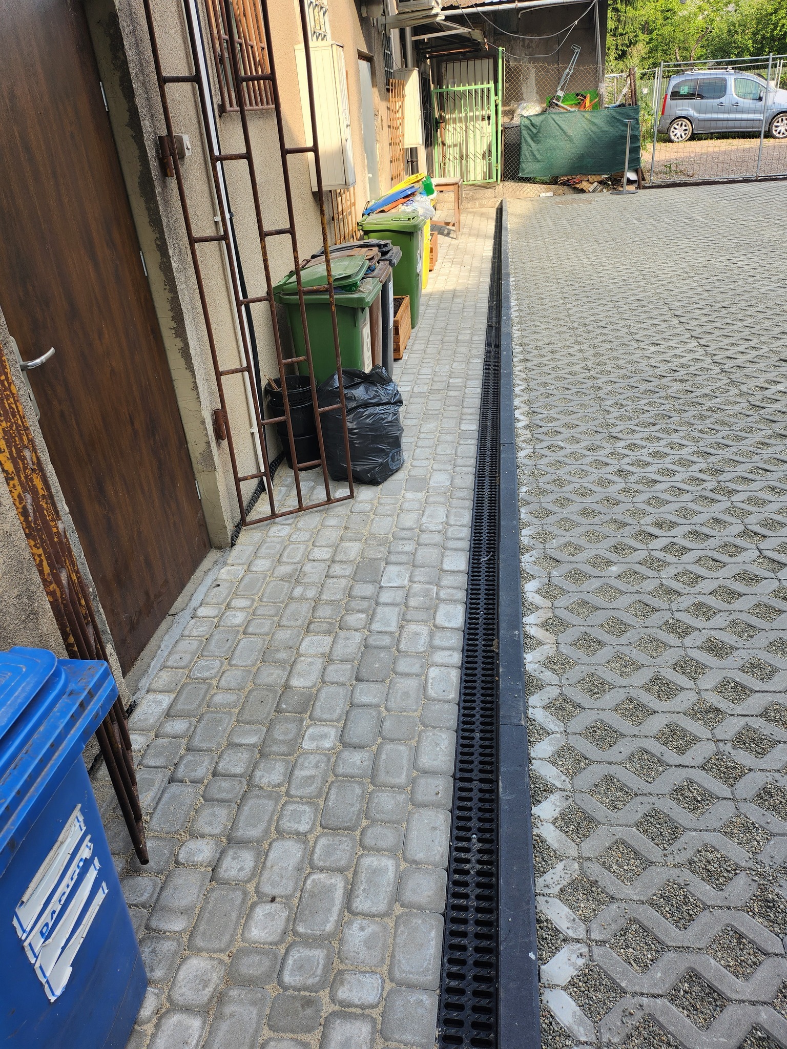 Szara kostka brukowa ułożona obok budynku z zamontowanym systemem drenażowym, widoczne pojemniki na śmieci i krawężnik z ażurowych płyt.