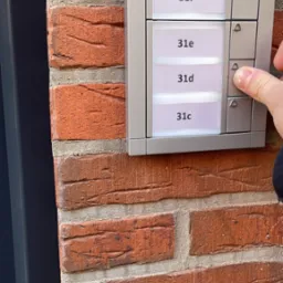 Dłoń naciska przycisk dzwonka w panelu domofonowym na ceglanej ścianie budynku z adresami 31c do 31h.