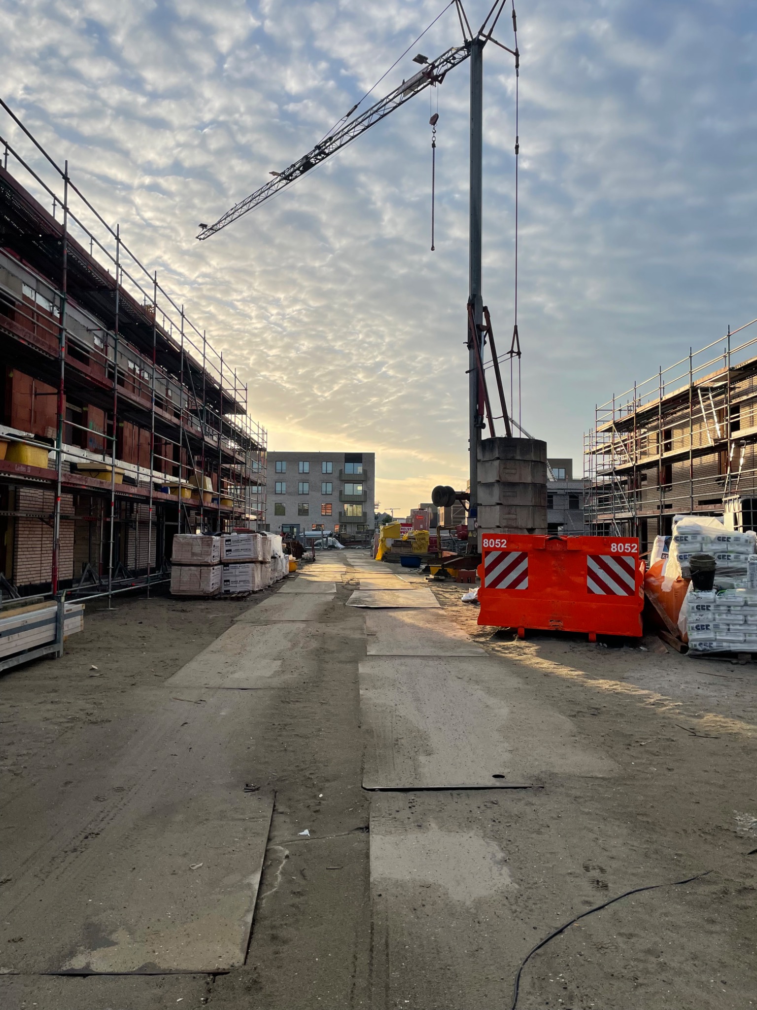 Plac budowy z rusztowaniami, cegłami, zaprawą, pomarańczowymi barierami z numerem 8052, dźwigiem i płytami betonowymi na ziemi w pochmurny dzień.