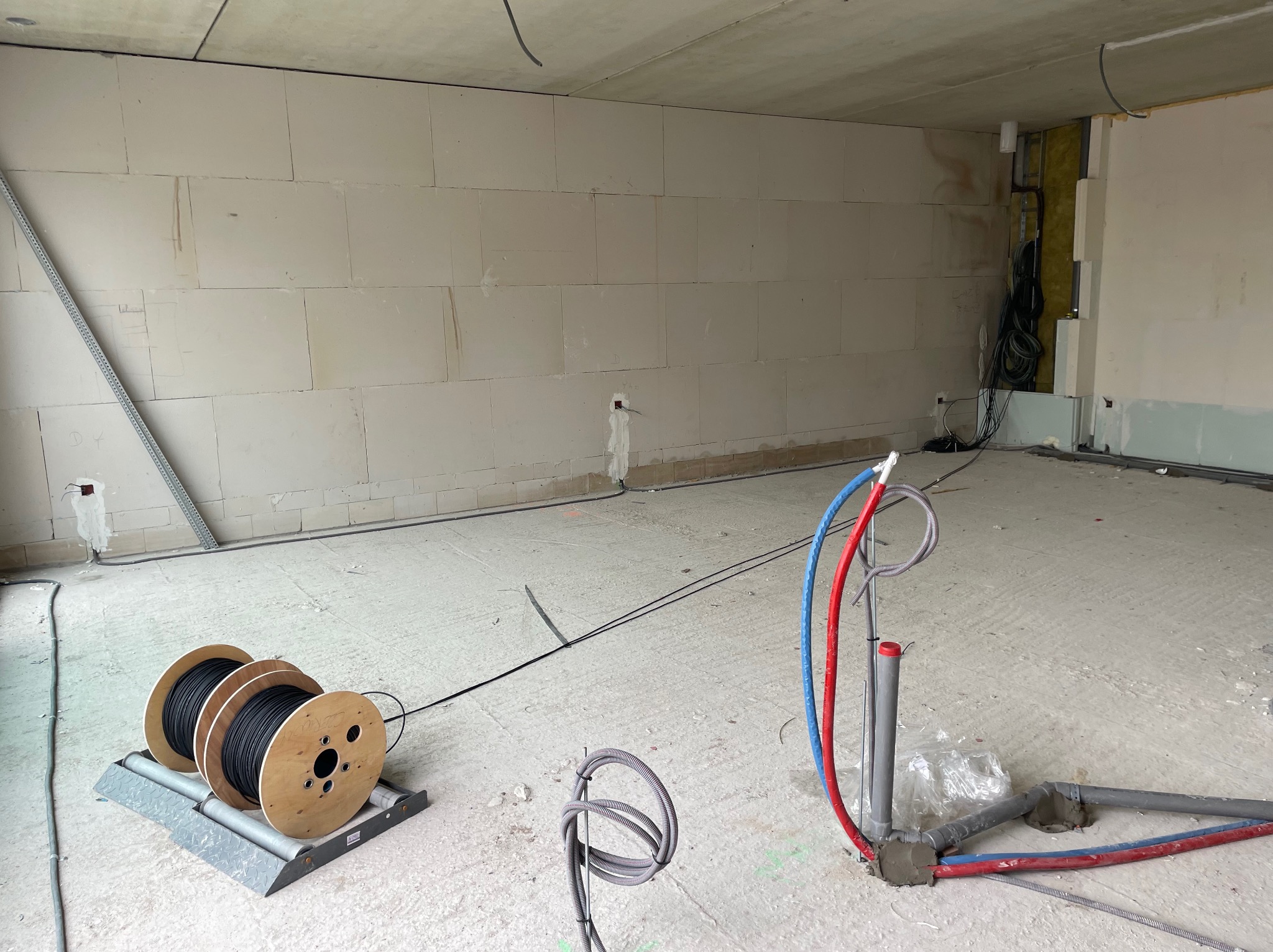 Wnętrze w trakcie prac: widoczne szpule z kablem na podajniku, rury instalacji CO w kolorach czerwonym i niebieskim oraz przewody elektryczne wystające ze ściany z betonu komórkowego.
