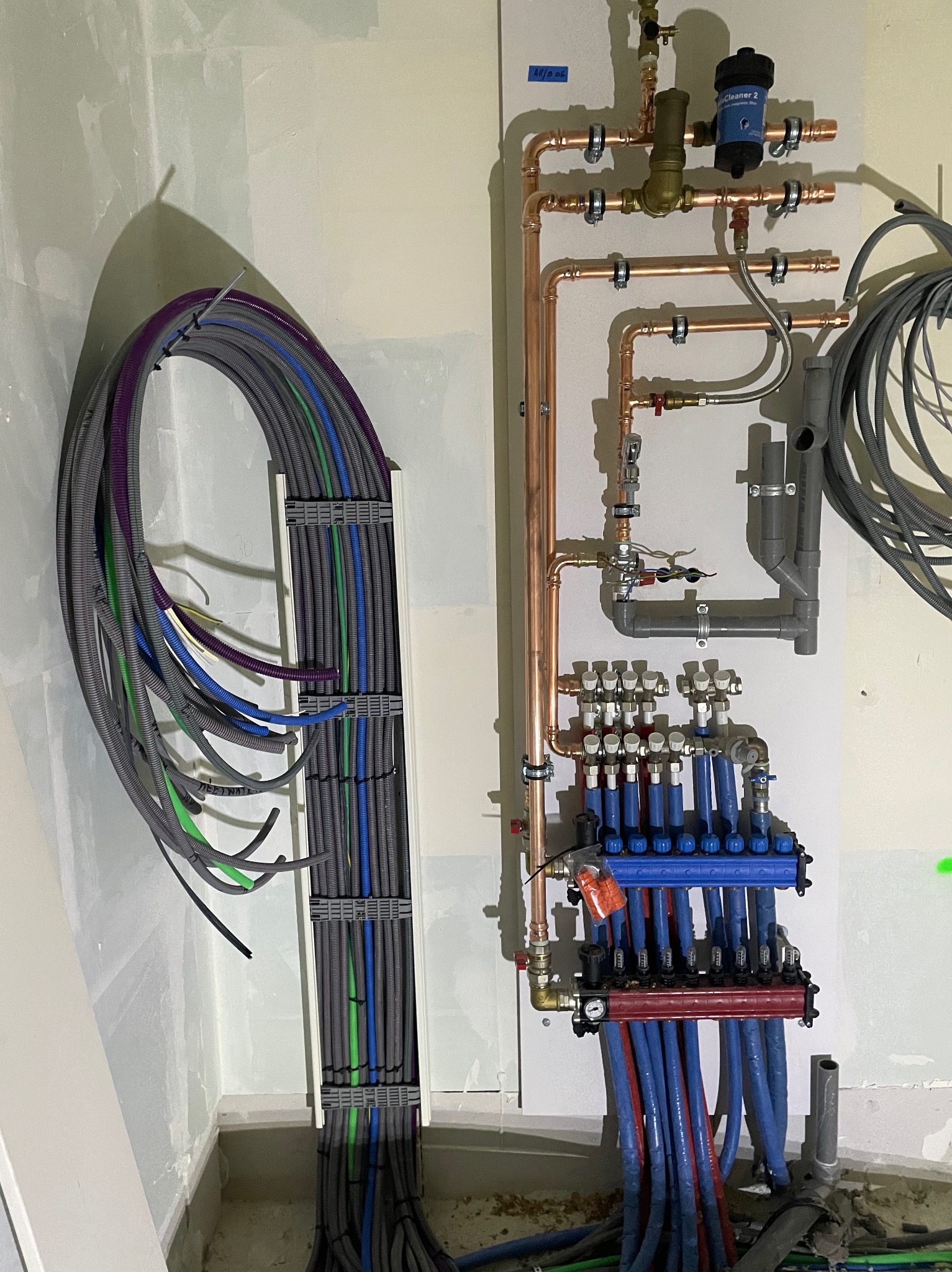 Rozdzielacz ogrzewania podłogowego z rurami w kolorach niebieskim i czerwonym, połączony z miedzianą instalacją, obok wiązka przewodów elektrycznych w korytku, ściany w stanie surowym.