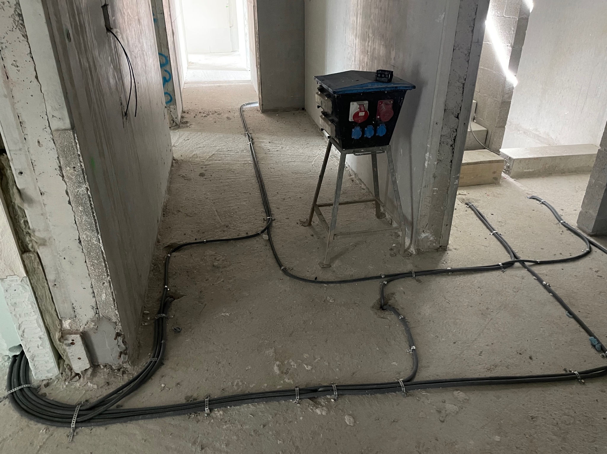 Rozprowadzone kable elektryczne w szarych peszlach, przymocowane metalowymi uchwytami do surowej betonowej podłogi w trakcie instalacji, z czarną skrzynką rozdzielczą na metalowym stojaku pośrodku...