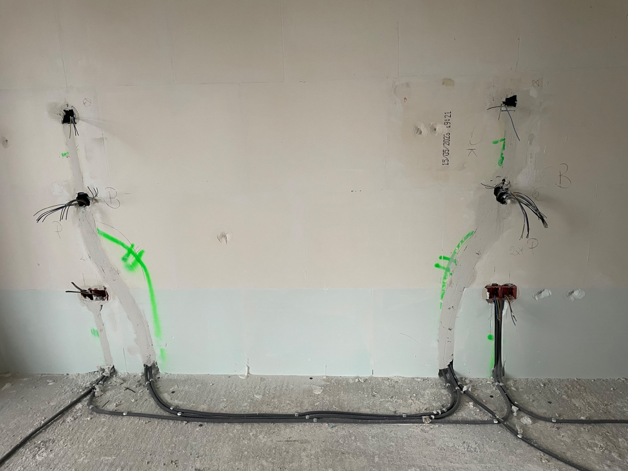 Ściana w trakcie prac elektrycznych: widoczne kable w peszlach, puszki instalacyjne, fragmenty tynku z zielonymi oznaczeniami i datą, surowy beton na podłodze.