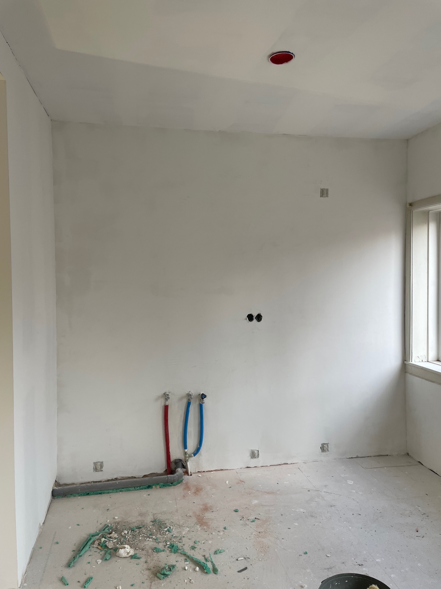 Surowe wnętrze w trakcie remontu: widoczne wystające rury wodne w kolorze czerwonym i niebieskim, szare rury kanalizacyjne z zieloną izolacją, otwory na gniazdka elektryczne w ścianie oraz resztki...