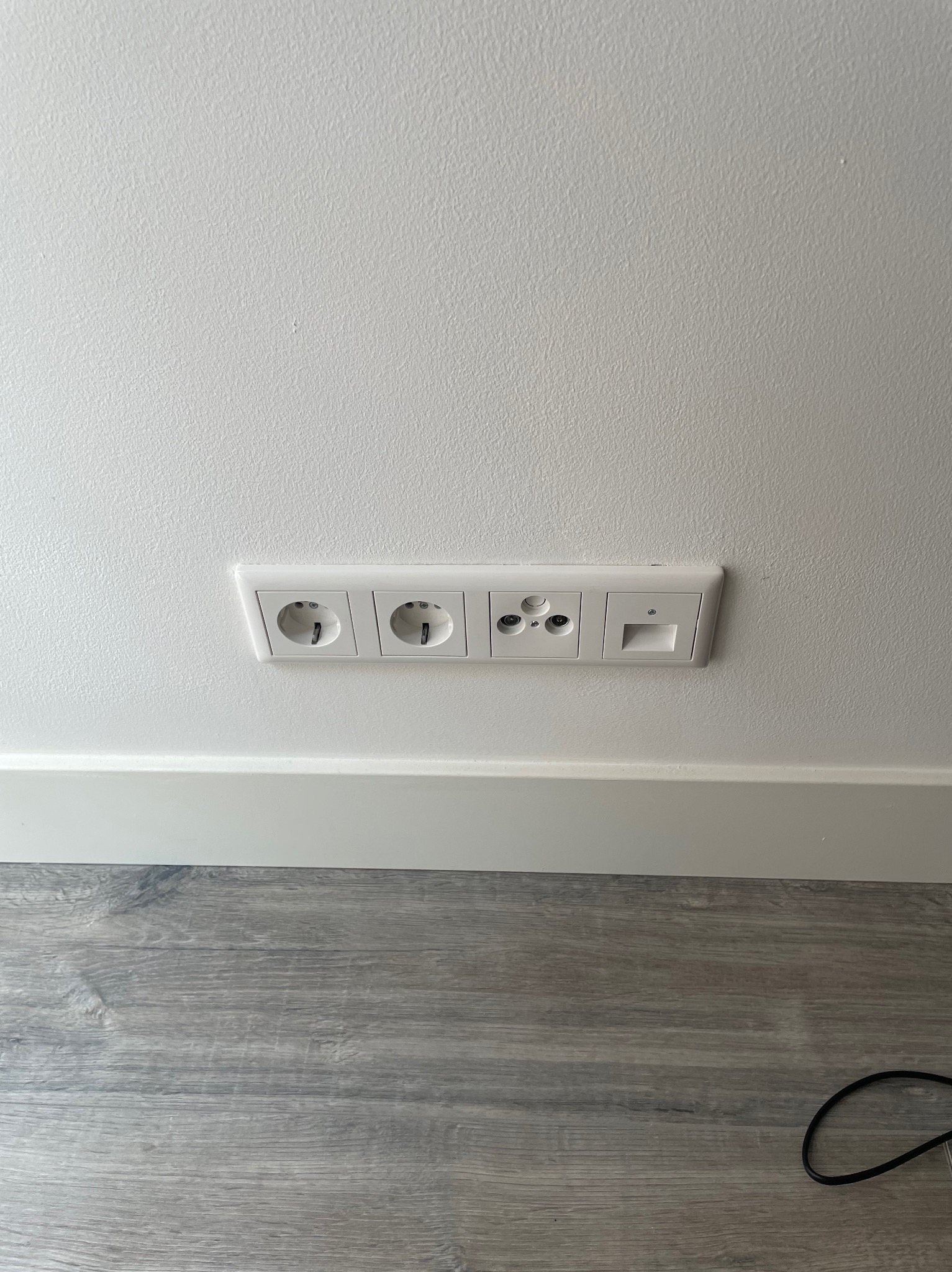 Gniazdka elektryczne i gniazdo antenowe w białej ramce, zamontowane w ścianie nad białą listwą przypodłogową i szarą podłogą imitującą drewno.