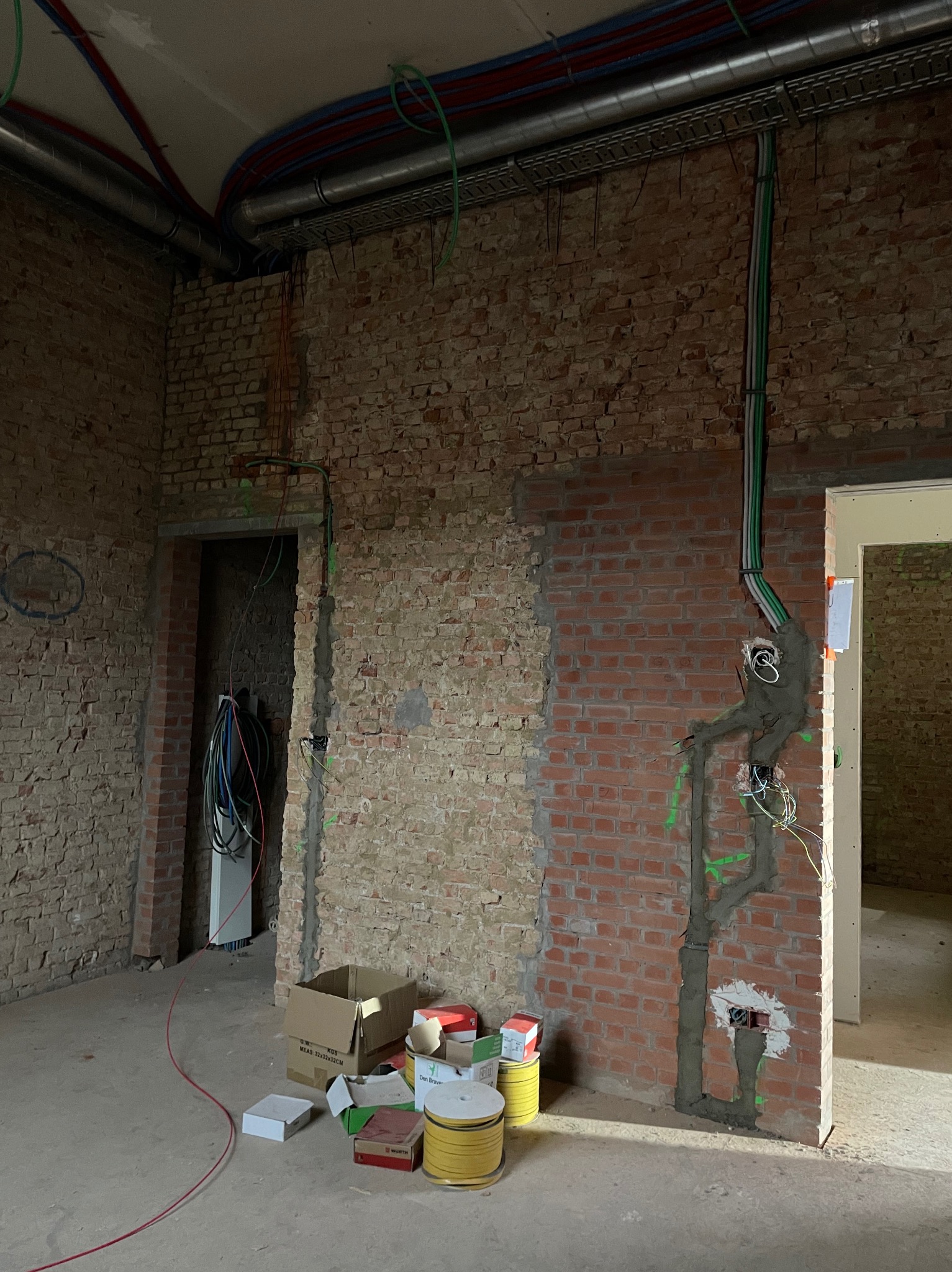 Instalacja elektryczna w surowym wnętrzu z ceglanymi ścianami: kable w peszlach prowadzone po ścianie, puszki elektryczne zamurowane, widoczne rury wentylacyjne pod sufitem i rozłożone materiały...