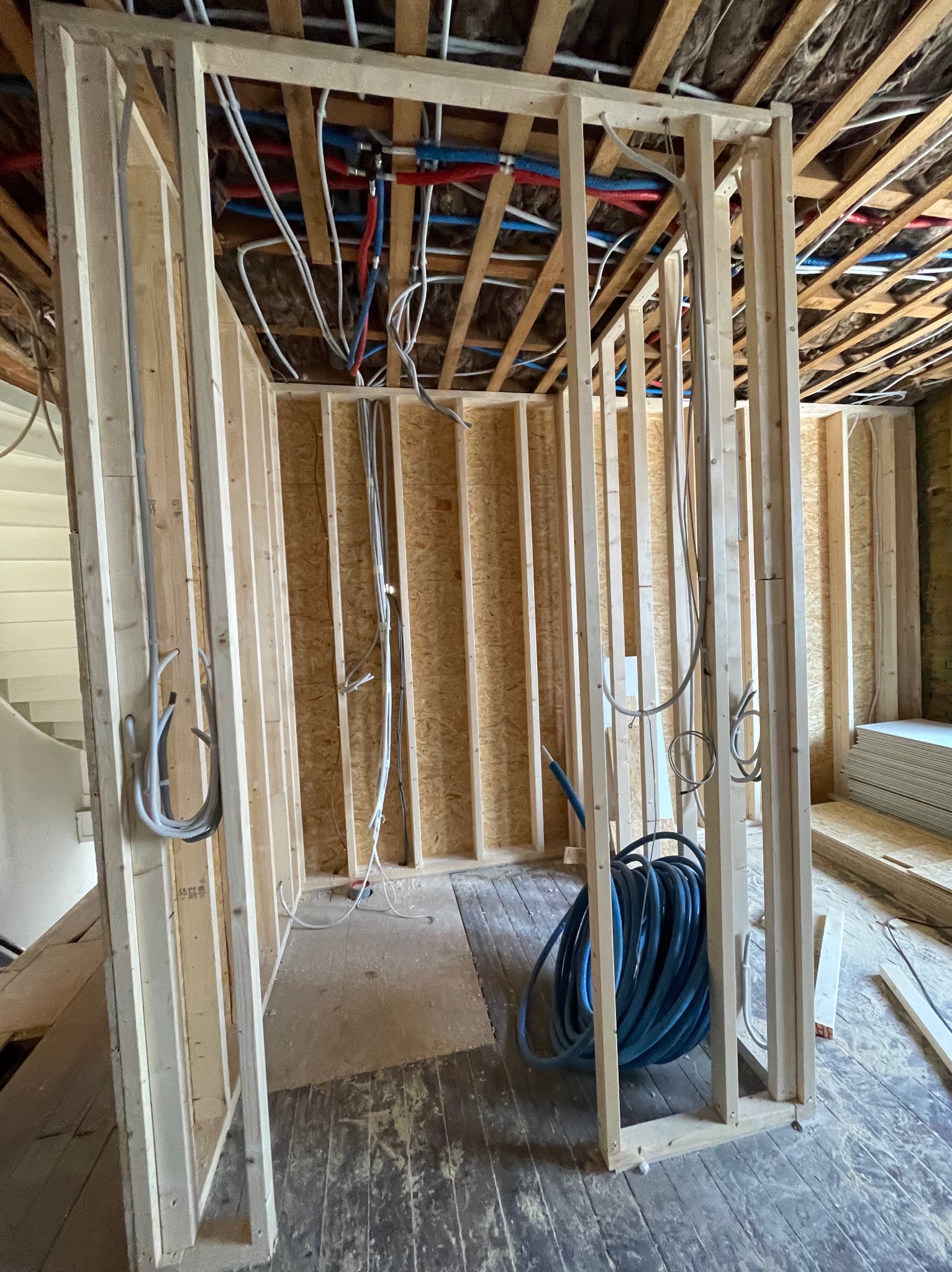 Surowa drewniana rama nowej ściany w trakcie budowy z widocznymi przewodami elektrycznymi i rurami instalacji wodnej pod sufitem