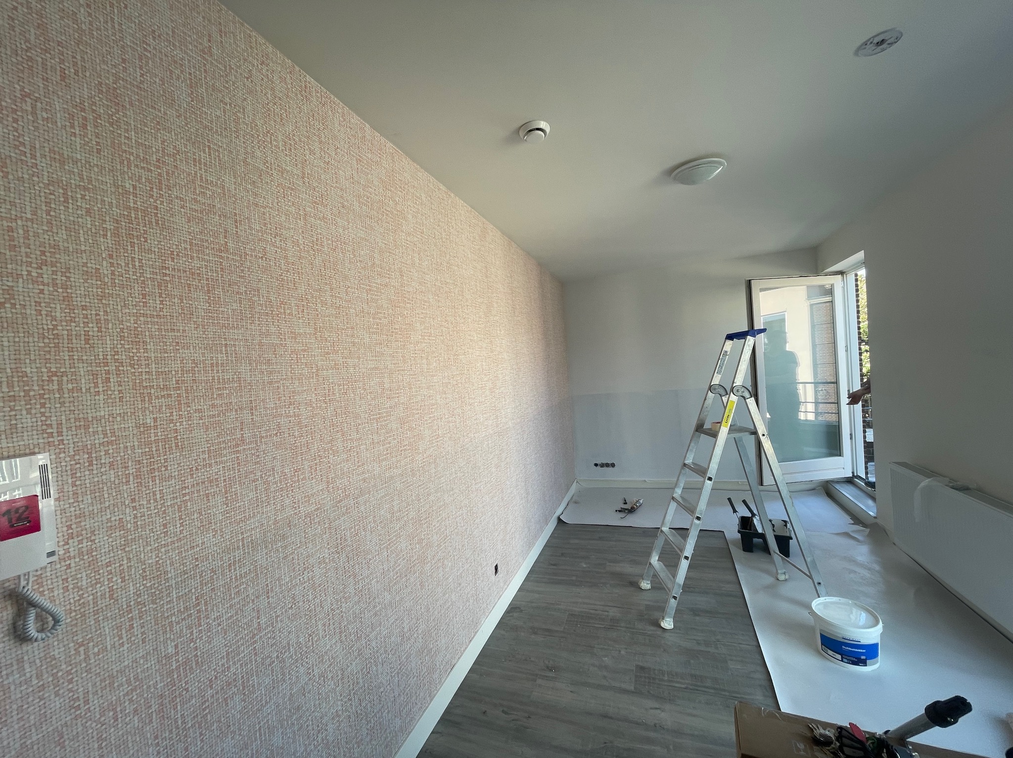 Pokój w trakcie remontu, ściana z tapetą o wzorze drobnej mozaiki, drabina aluminiowa, wiadro z farbą i narzędzia malarskie na podłodze zabezpieczonej folią malarską, widoczny domofon na ścianie.