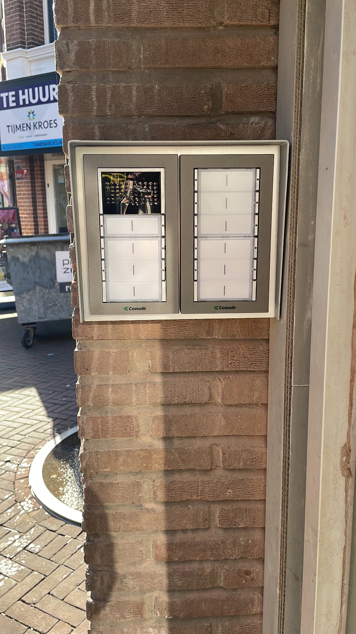 Zamontowany na ceglanej ścianie zewnętrznej dwupanelowy panel domofonowy Comelit z jednym panelem zawierającym wizytówki i drugim z kamerą i przyciskami dzwonka.