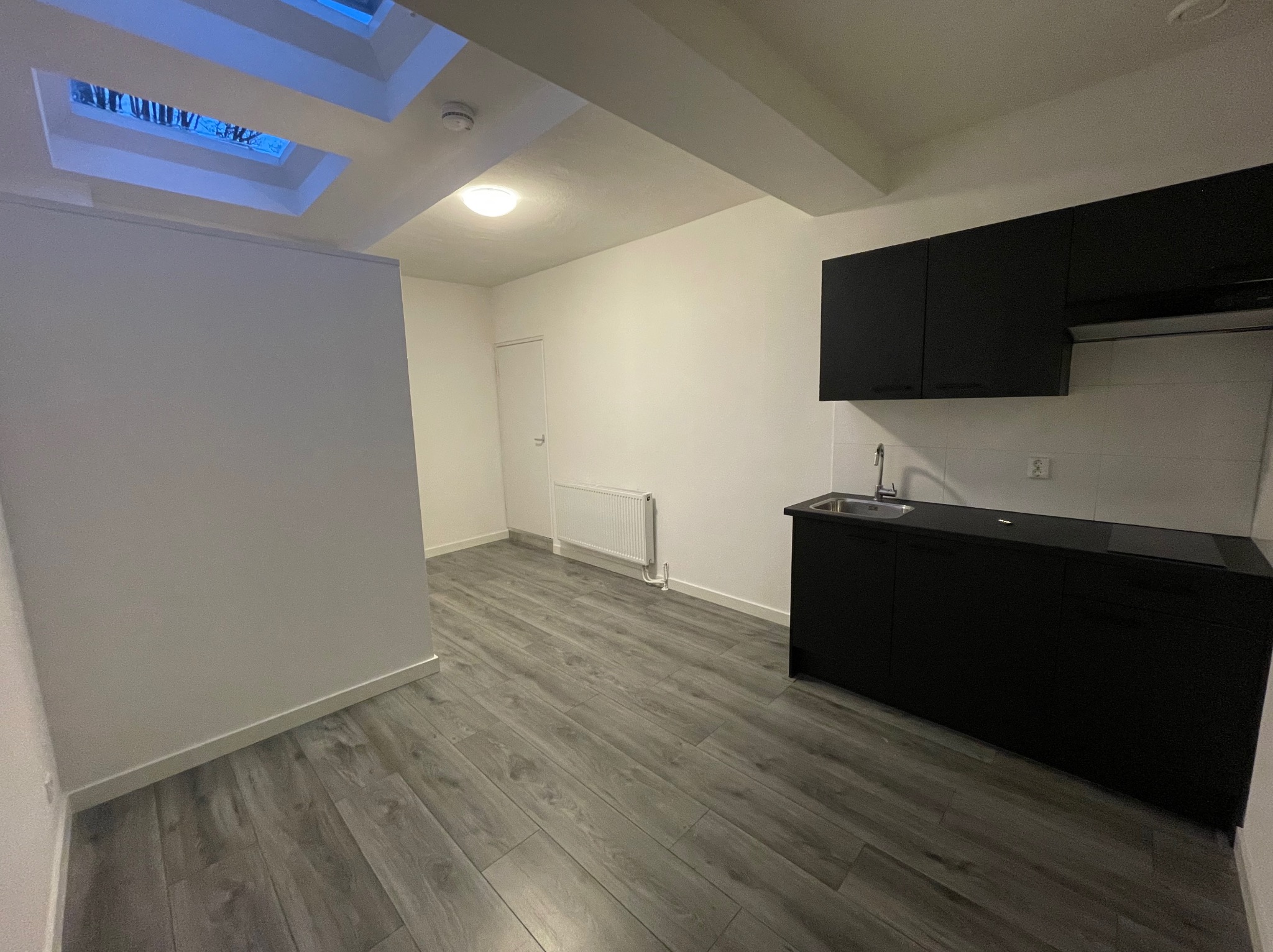 Wnętrze kuchni z czarnymi szafkami i szarą podłogą imitującą drewno, oświetlone światłem dziennym wpadającym przez okno dachowe.