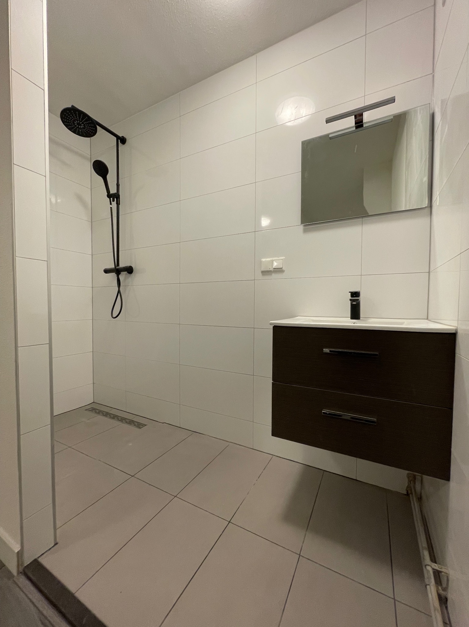 Nowoczesna łazienka z czarną armaturą: deszczownica, bateria umywalkowa, szafka pod umywalką z dwiema szufladami, lustro z oświetleniem nad umywalką, białe płytki na ścianach i szare na podłodze.