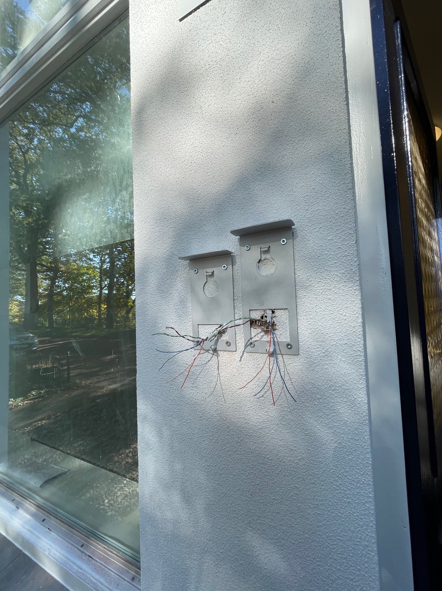 Dwa uchwyty montażowe domofonu/wideofonu przymocowane do białej, teksturowanej ściany zewnętrznej, z widocznymi przewodami elektrycznymi przed podłączeniem, obok okna z odbijającym się krajobrazem.