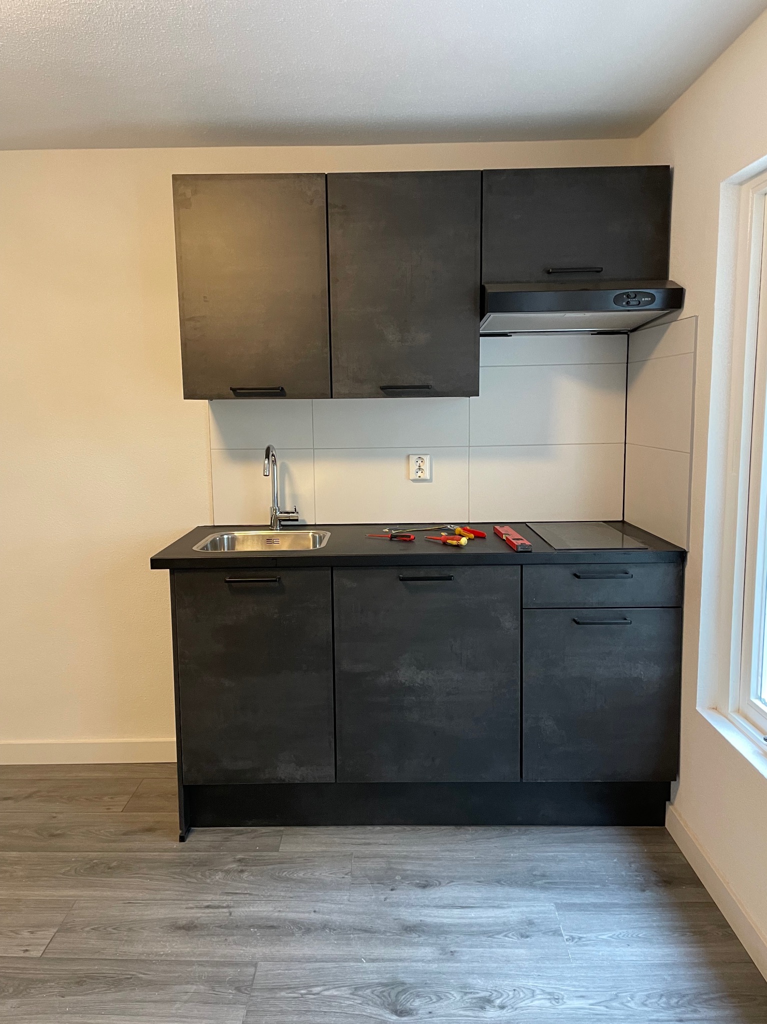 Czarna zabudowa kuchenna z płytą indukcyjną, zlewozmywakiem i okapem, widoczne narzędzia na blacie, gniazdko elektryczne na ścianie.