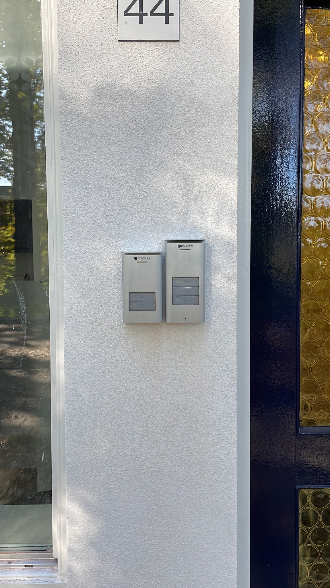 Dwa panele domofonowe Smartwares zamontowane na białej elewacji budynku obok drzwi z szybami w odcieniach bursztynu, widoczny numer domu 44.