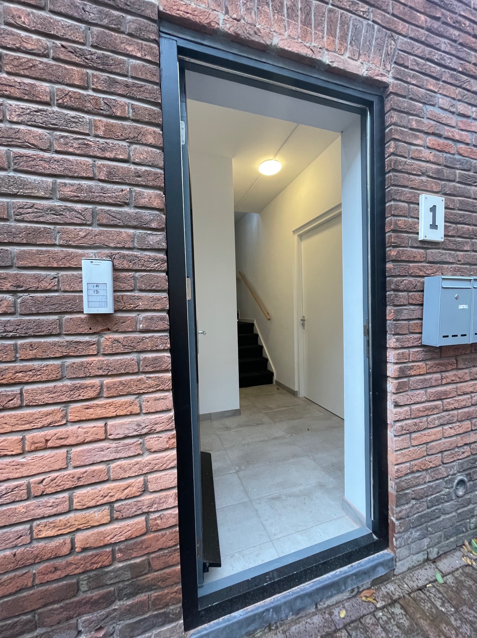 Wejście do budynku z cegły z domofonem i skrzynkami pocztowymi, widok na klatkę schodową z czarnymi schodami i białymi ścianami.